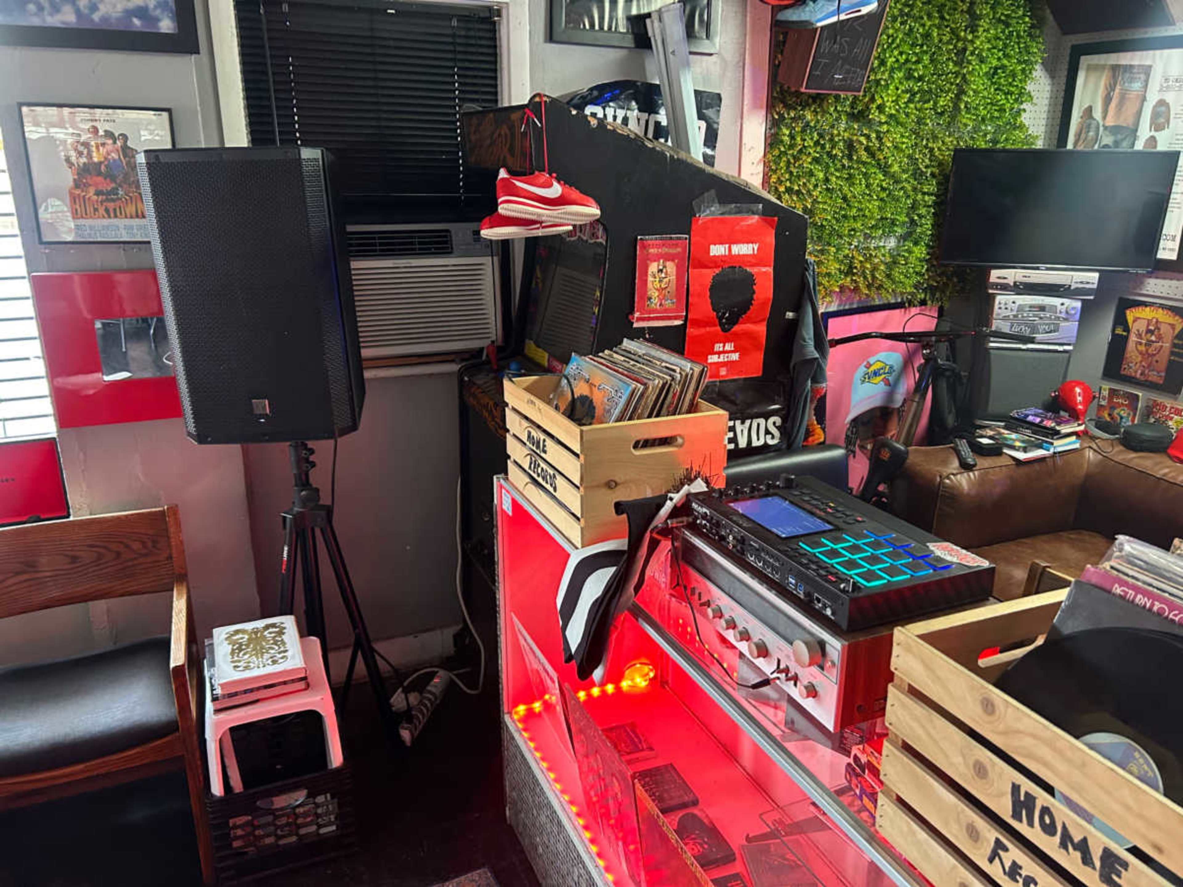 The image shows a small music setup featuring a turntable, speakers, and vinyl records displayed in crates, with a cozy seating area and decorative wall elements.
