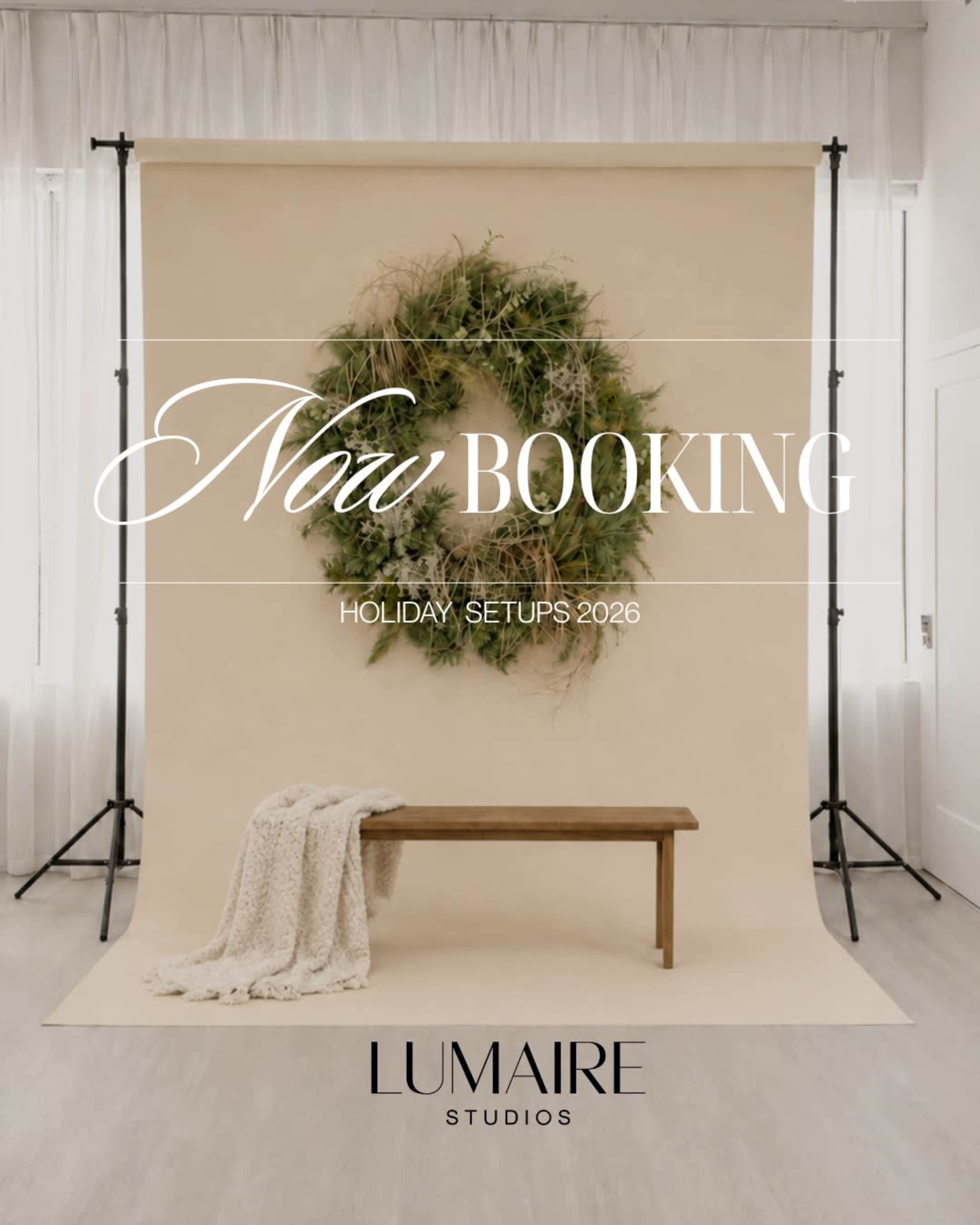 A simple photo studio setup features a beige backdrop with a large wreath, a wooden bench in front, and a cozy blanket draped beside it.