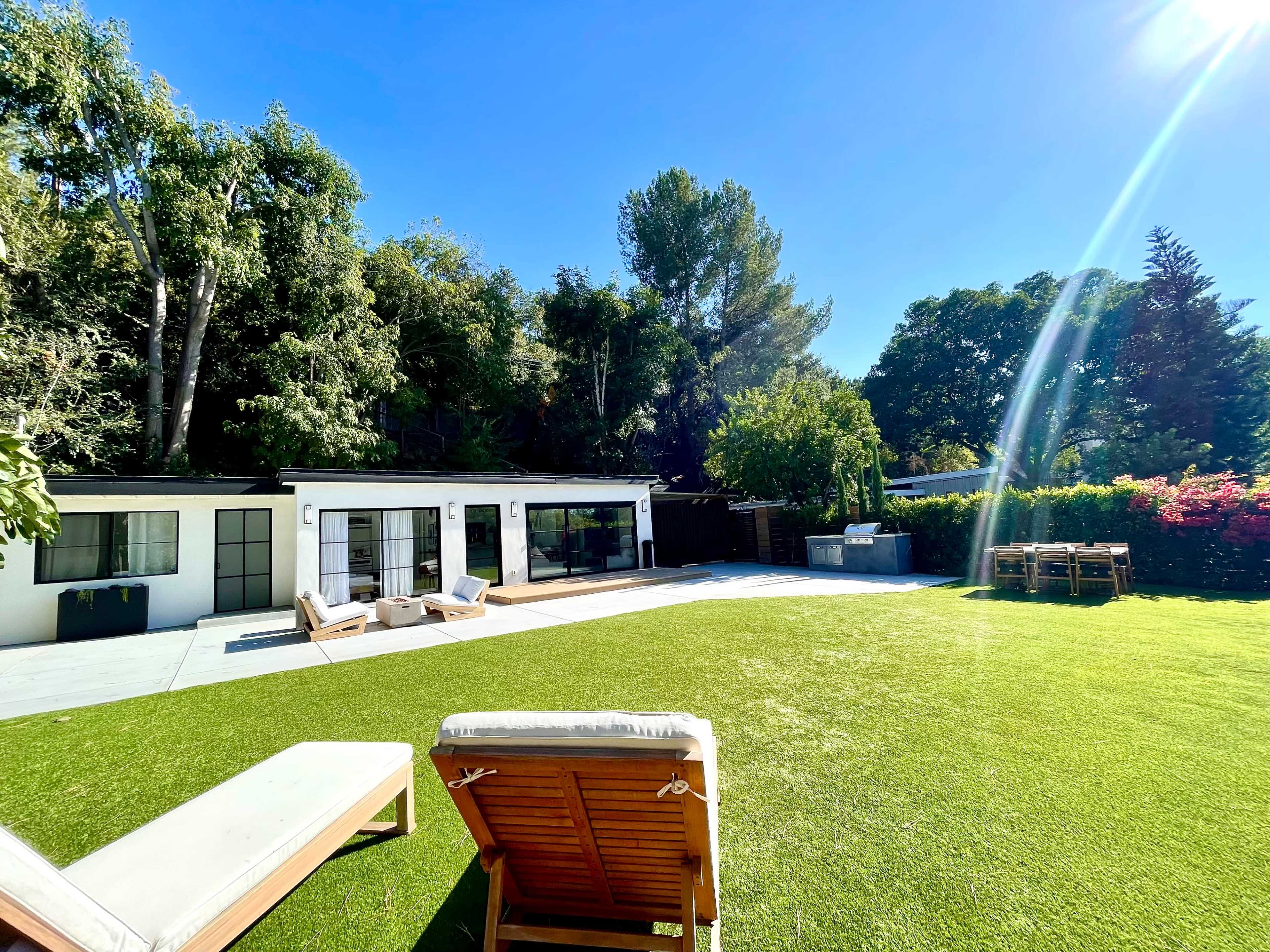 A spacious backyard features a green lawn, a modern house with large windows, and outdoor seating areas.