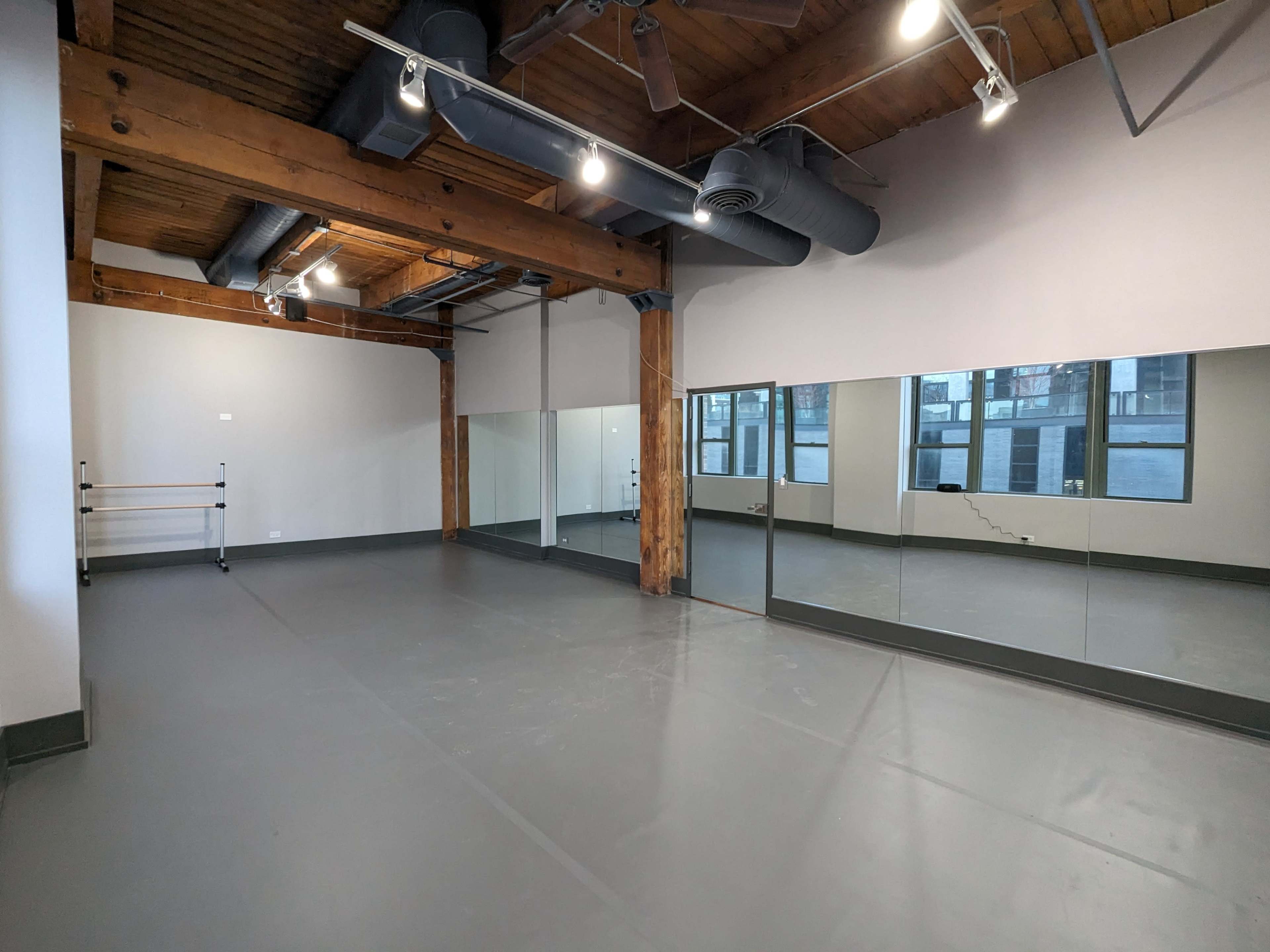 A spacious room with wooden beams and large mirrors along one wall, featuring a gray floor and ample natural light.
