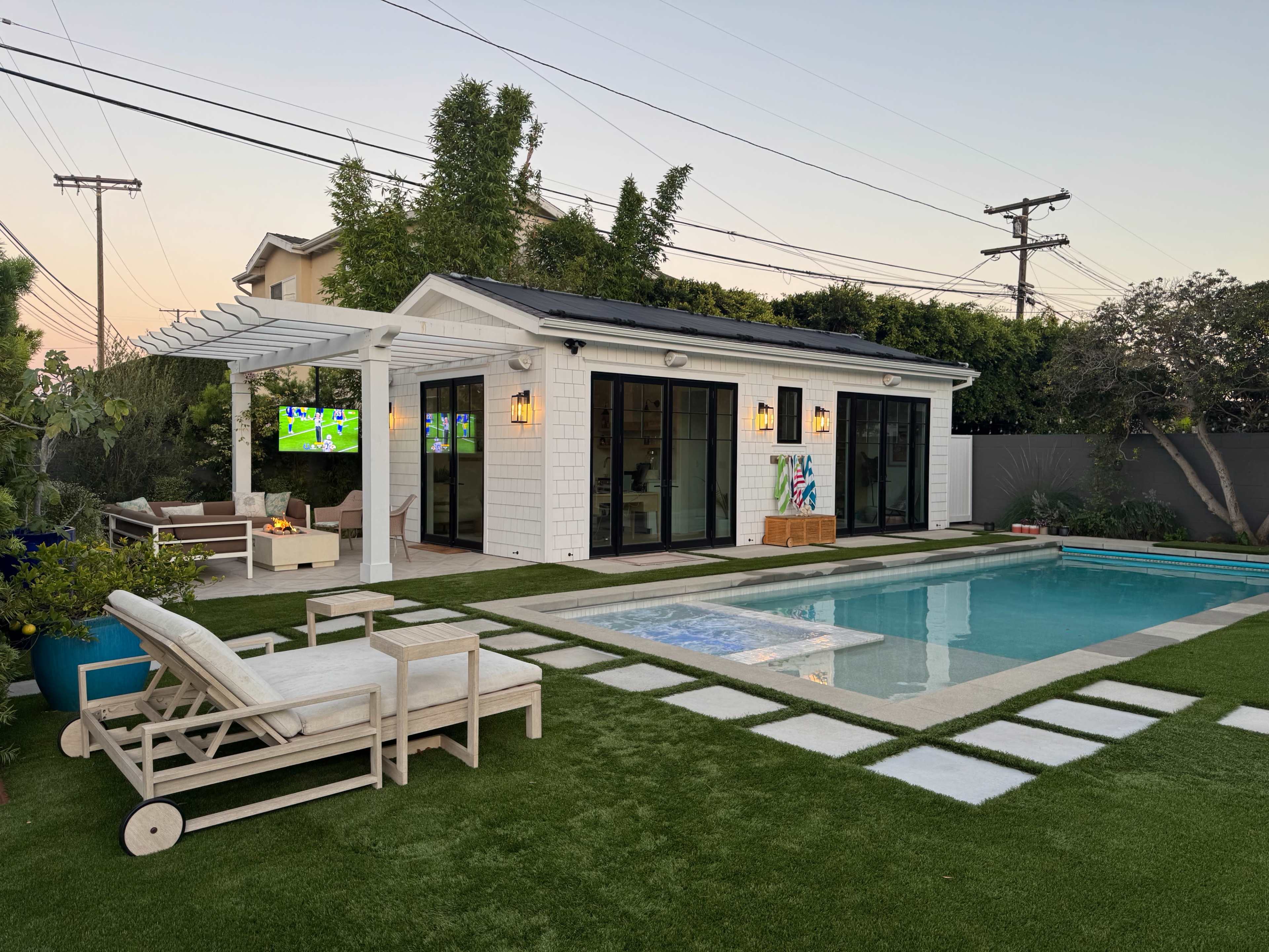 A modern backyard features a swimming pool, a small guest house with large glass doors, and outdoor seating areas on green grass.