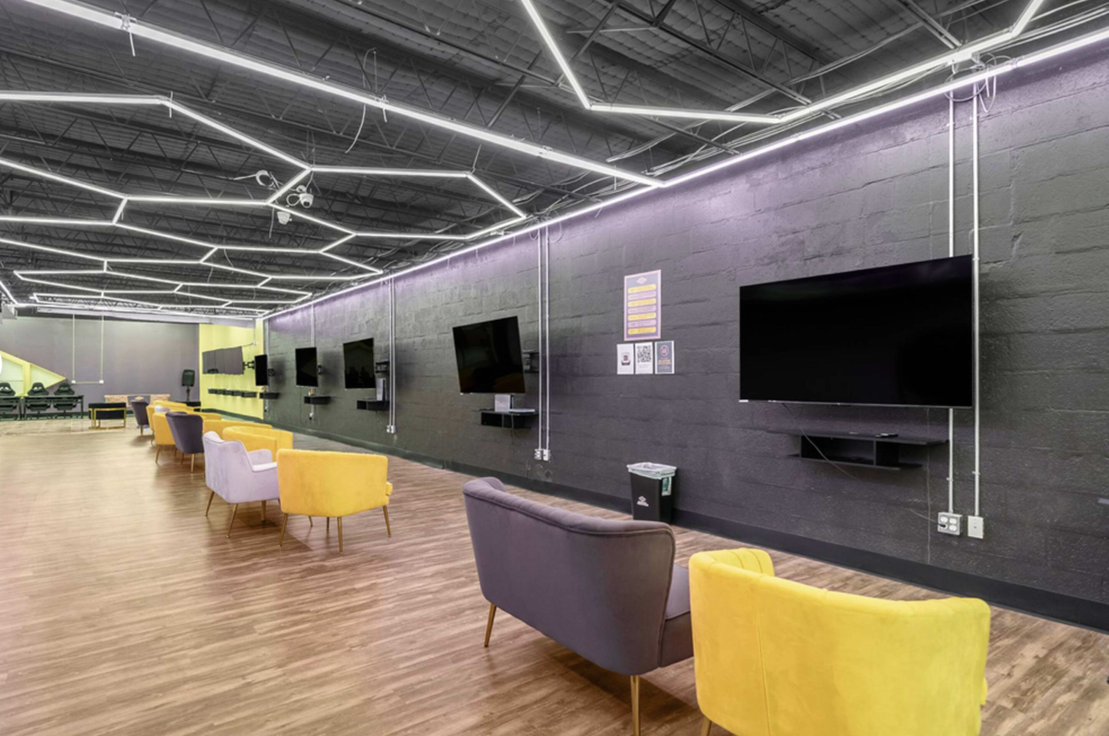 The scene features a modern lounge area with colorful chairs and wall-mounted televisions against dark walls, illuminated by geometric light fixtures overhead.