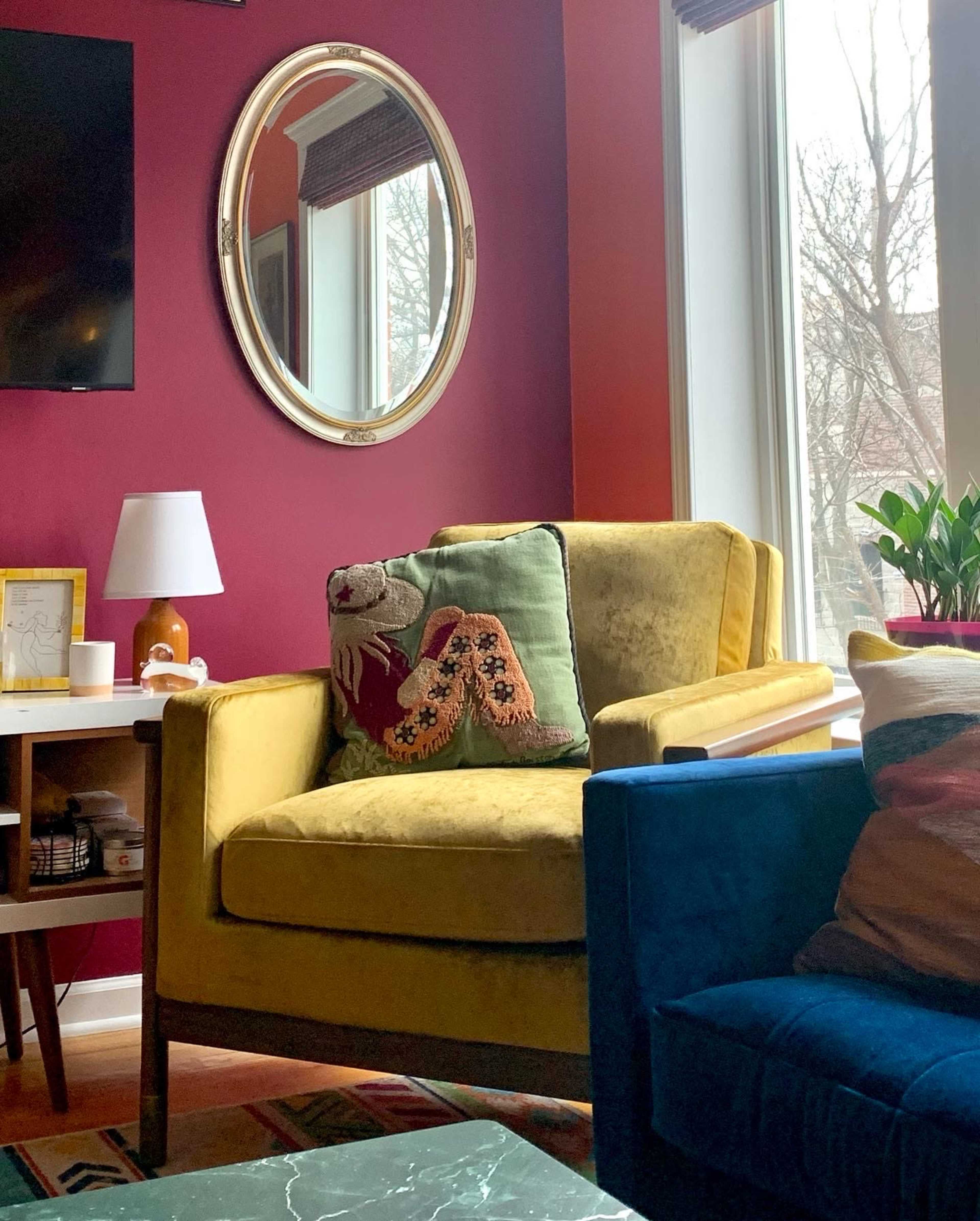 A yellow armchair with a decorative pillow is positioned in a room featuring a maroon wall and large windows, along with a lamp and a plant nearby.