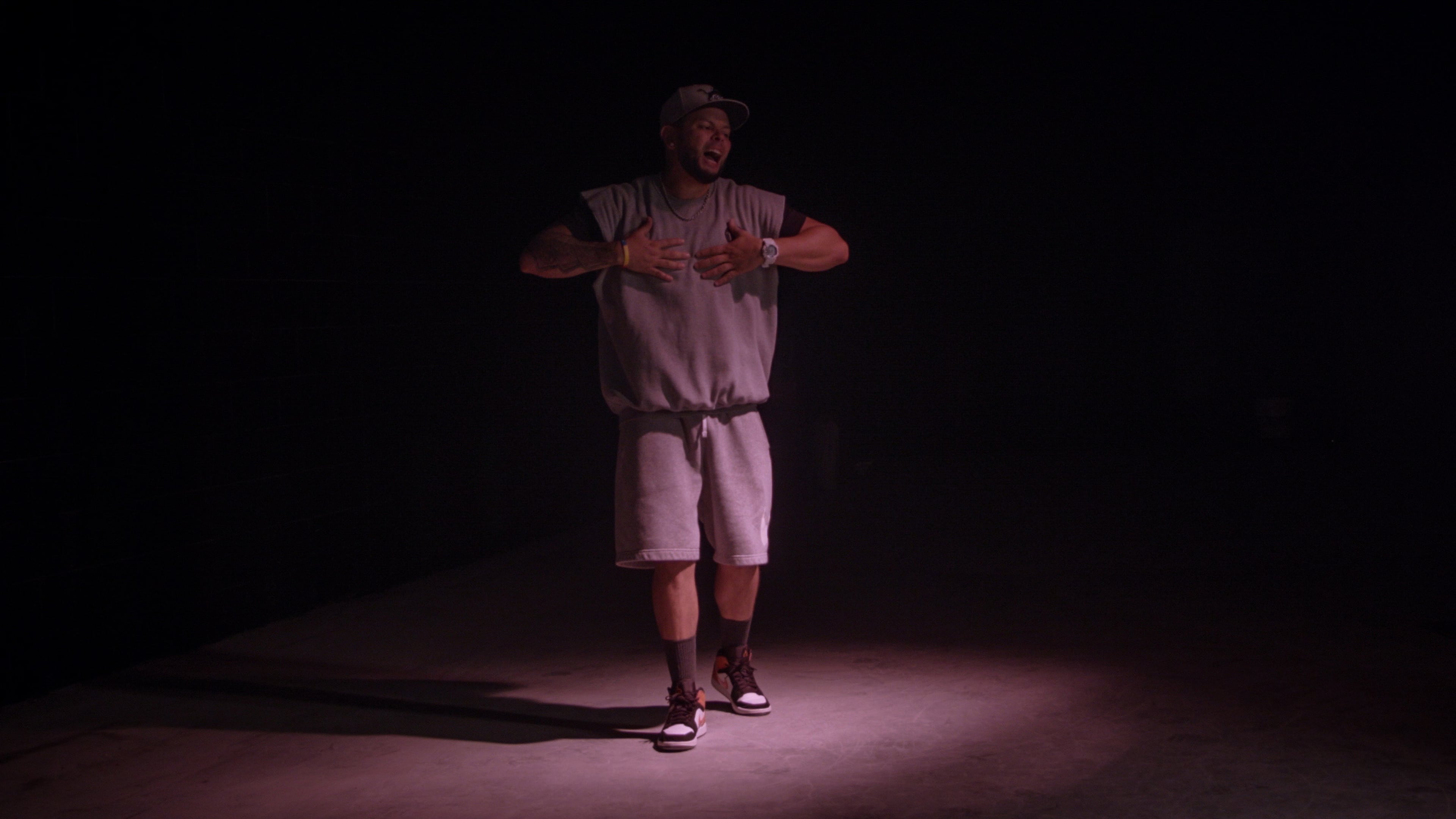 A performer stands in a dimly lit space, wearing a gray outfit and a cap, with hands clasped near their chest.