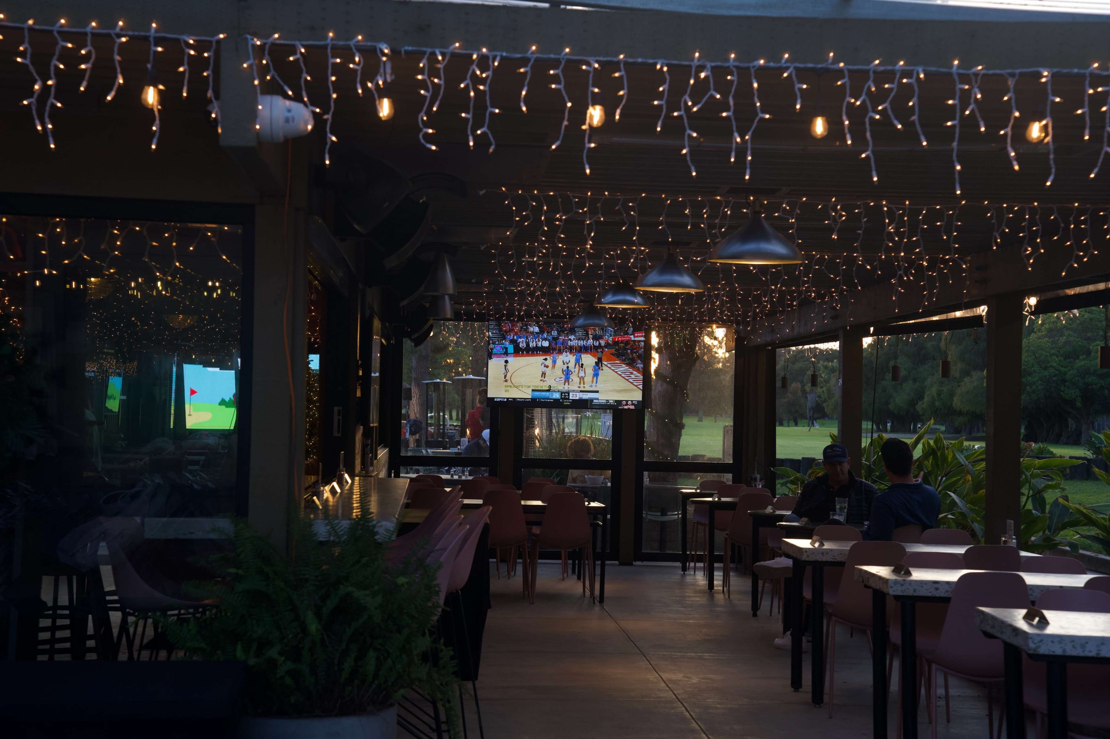 The image shows a dimly lit outdoor dining area with string lights overhead and a television mounted on the wall displaying a sports game.