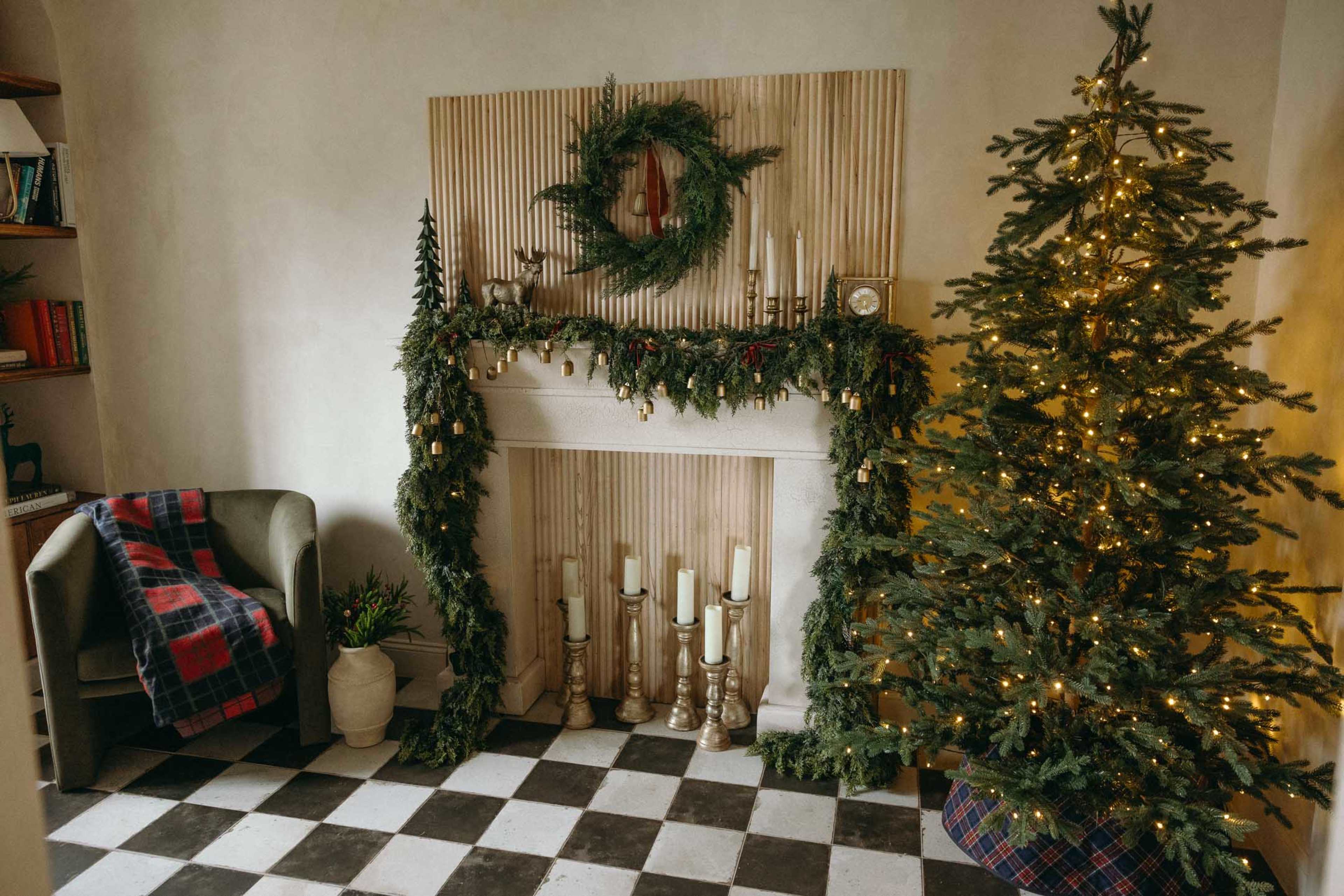 The image shows a cozy room decorated for the holidays, featuring a Christmas tree, a fireplace adorned with garlands, and a plaid blanket draped over a green armchair.