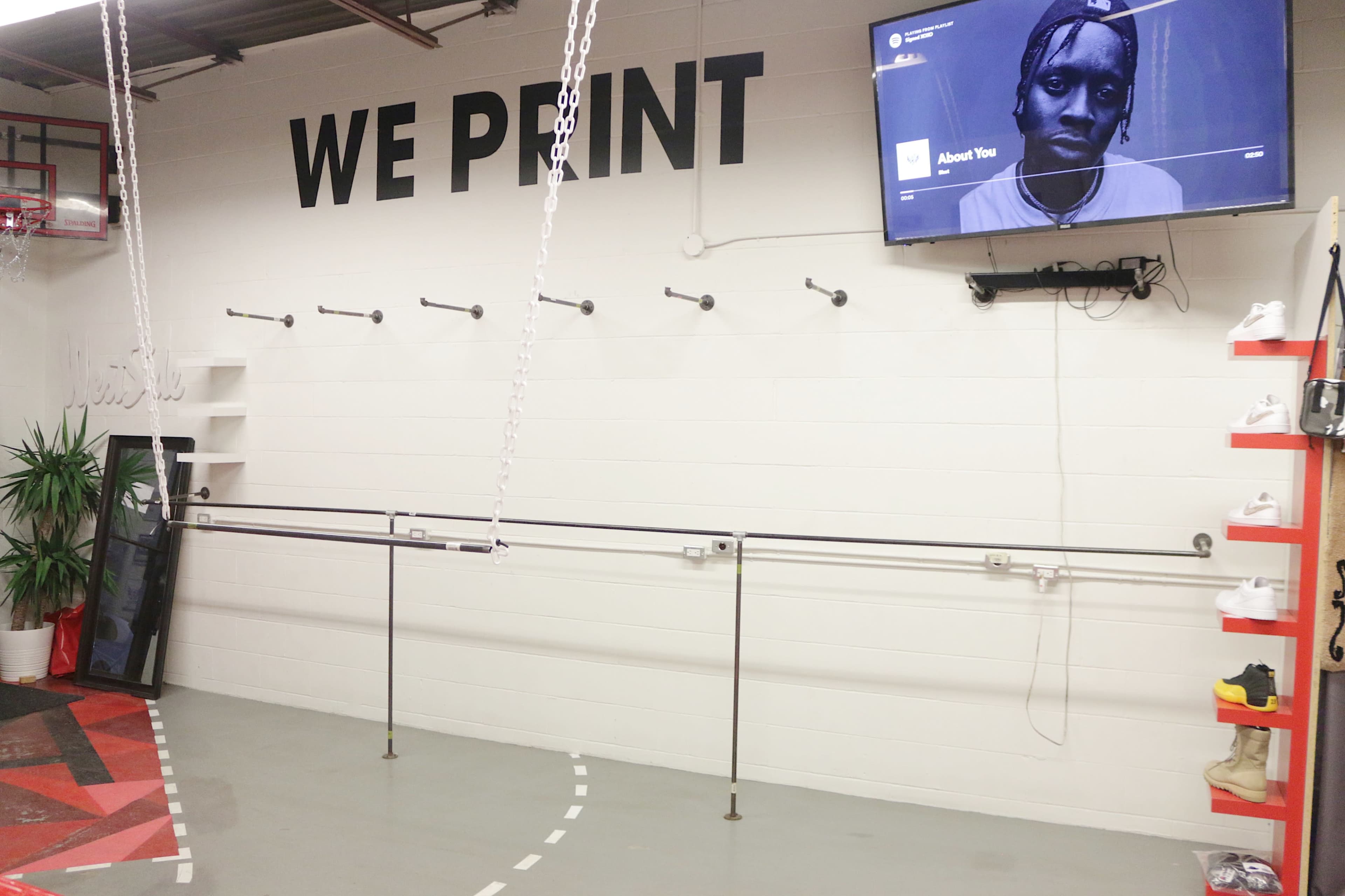 An interior space featuring a wall with the phrase "WE PRINT," a television screen mounted on the wall, and a row of hooks and shelves along one side.