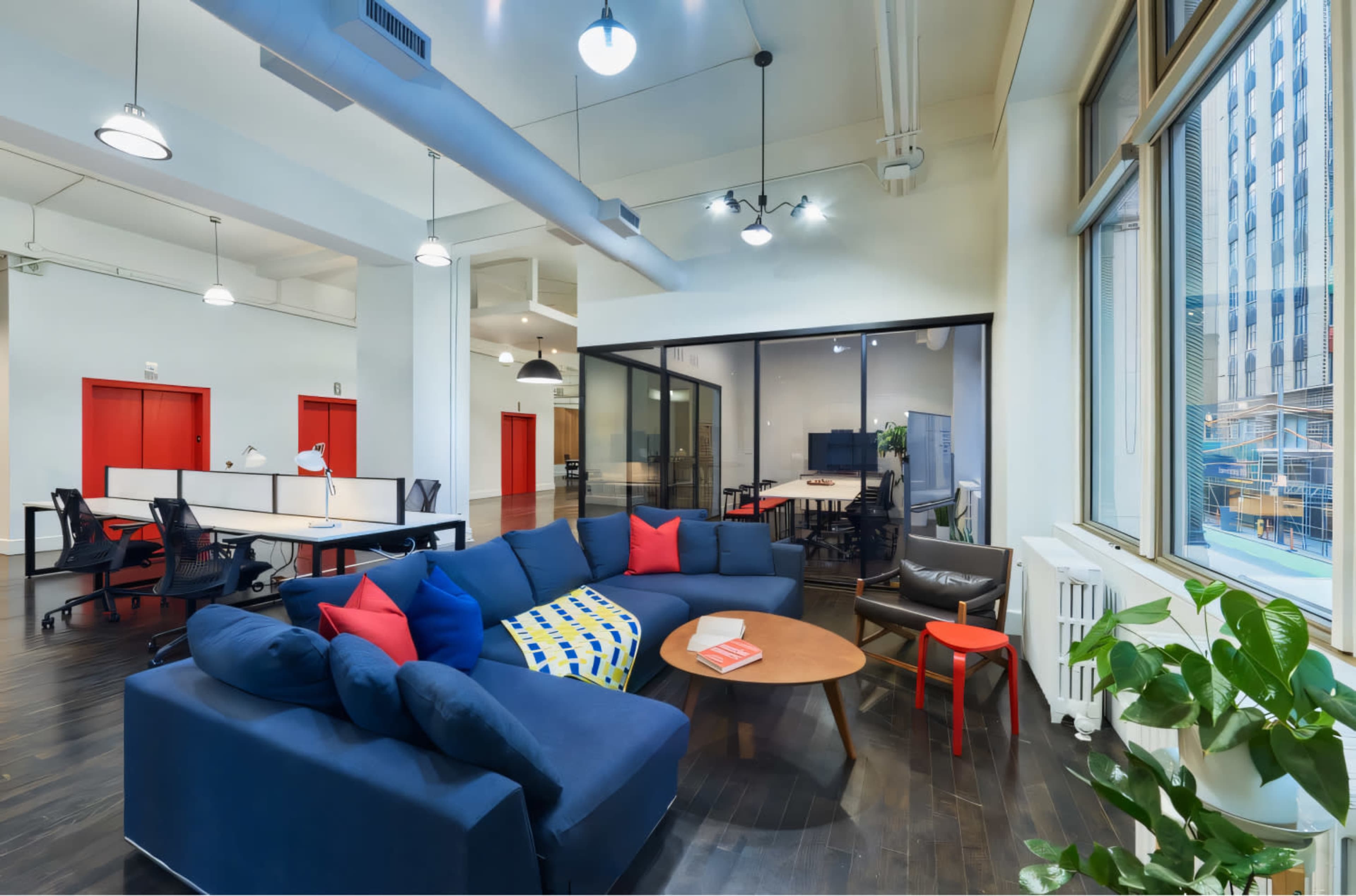 A modern office space featuring a blue sectional sofa with colorful pillows, a coffee table, and workstations with black office chairs.