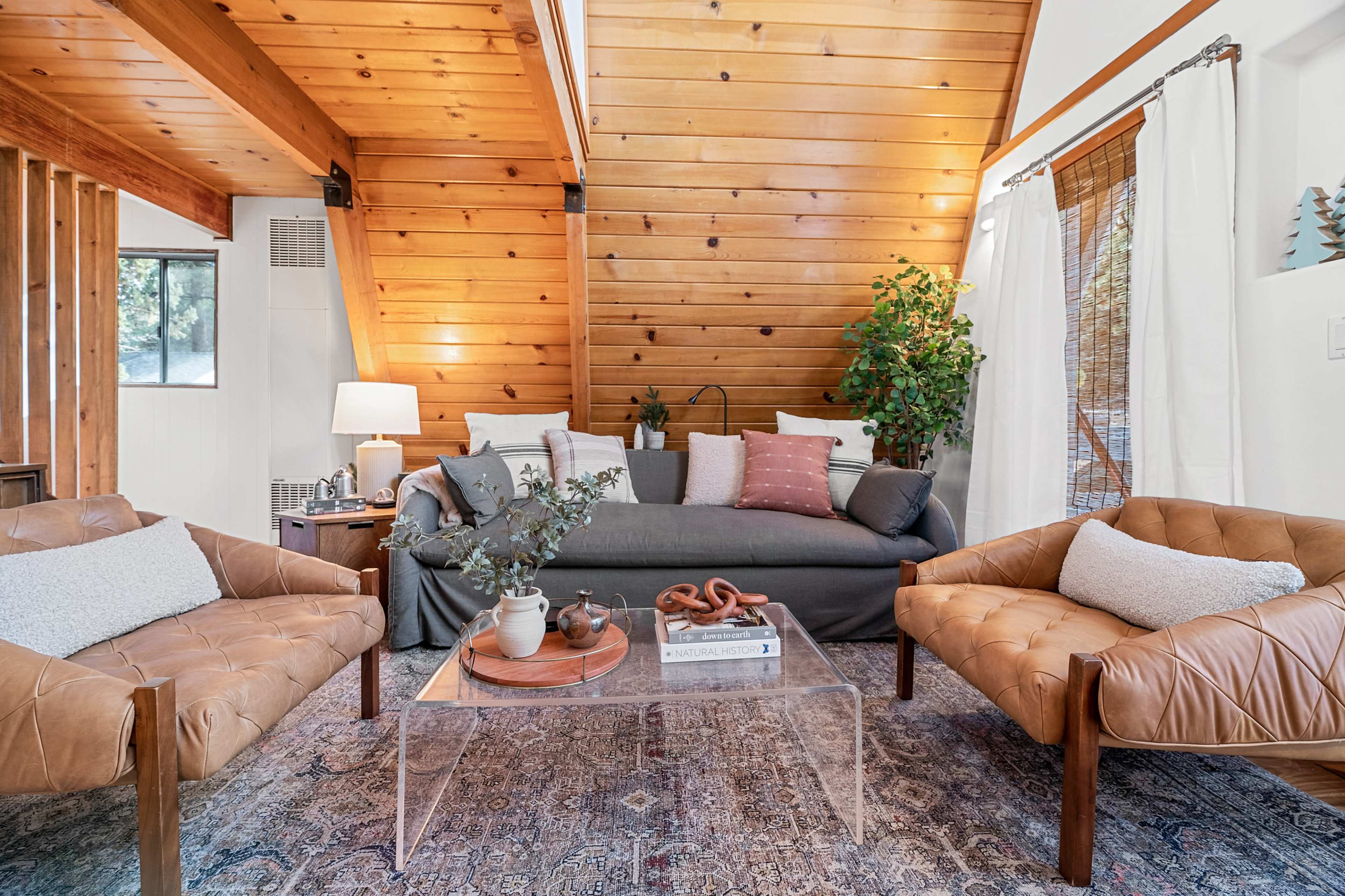 The cozy living room features a gray sofa, two tan leather chairs, a glass coffee table, and wooden beams on the ceiling.