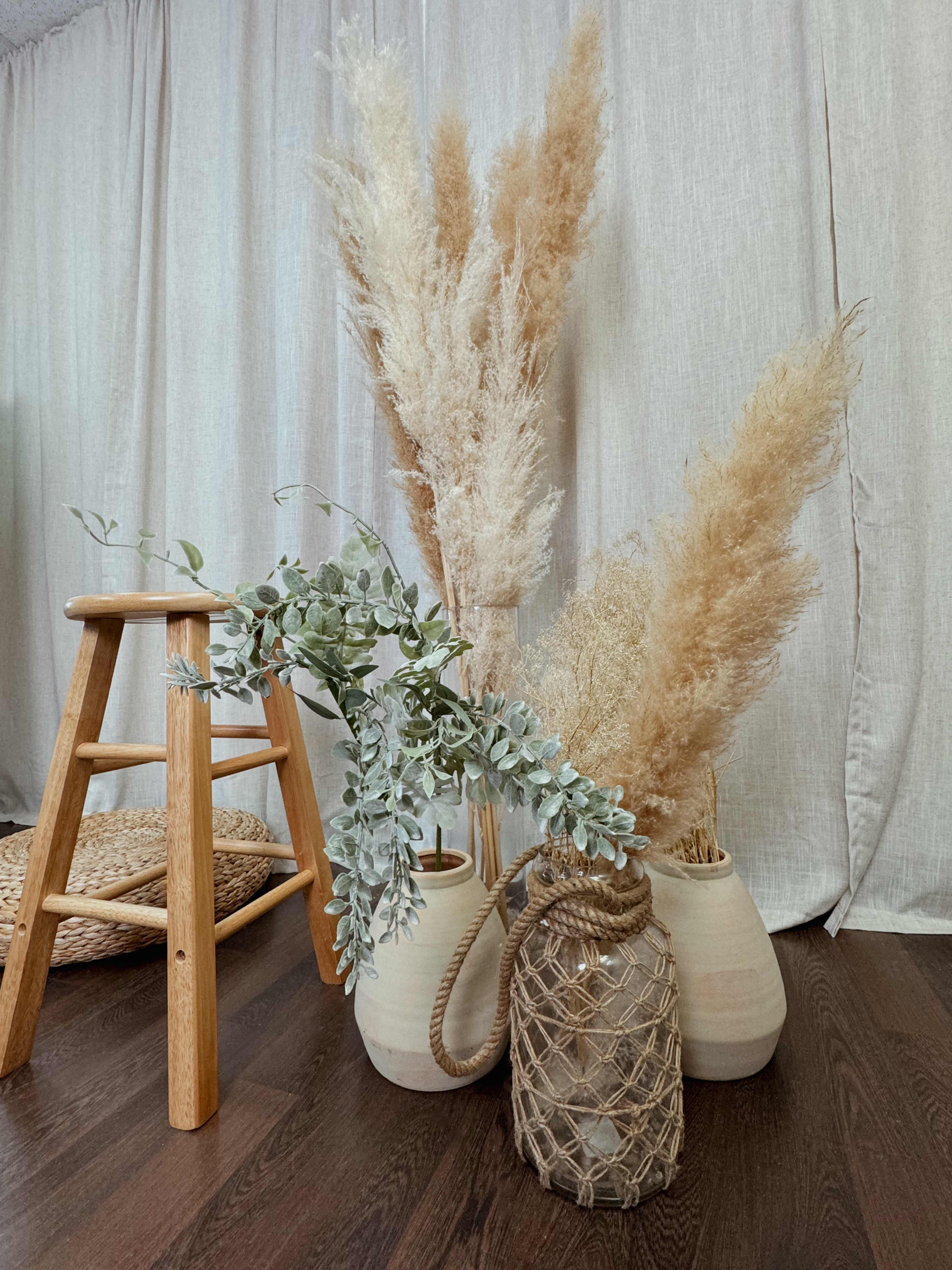 A wooden stool stands beside two large, decorated vases filled with dried pampas grass and greenery, set against a light-colored fabric backdrop.