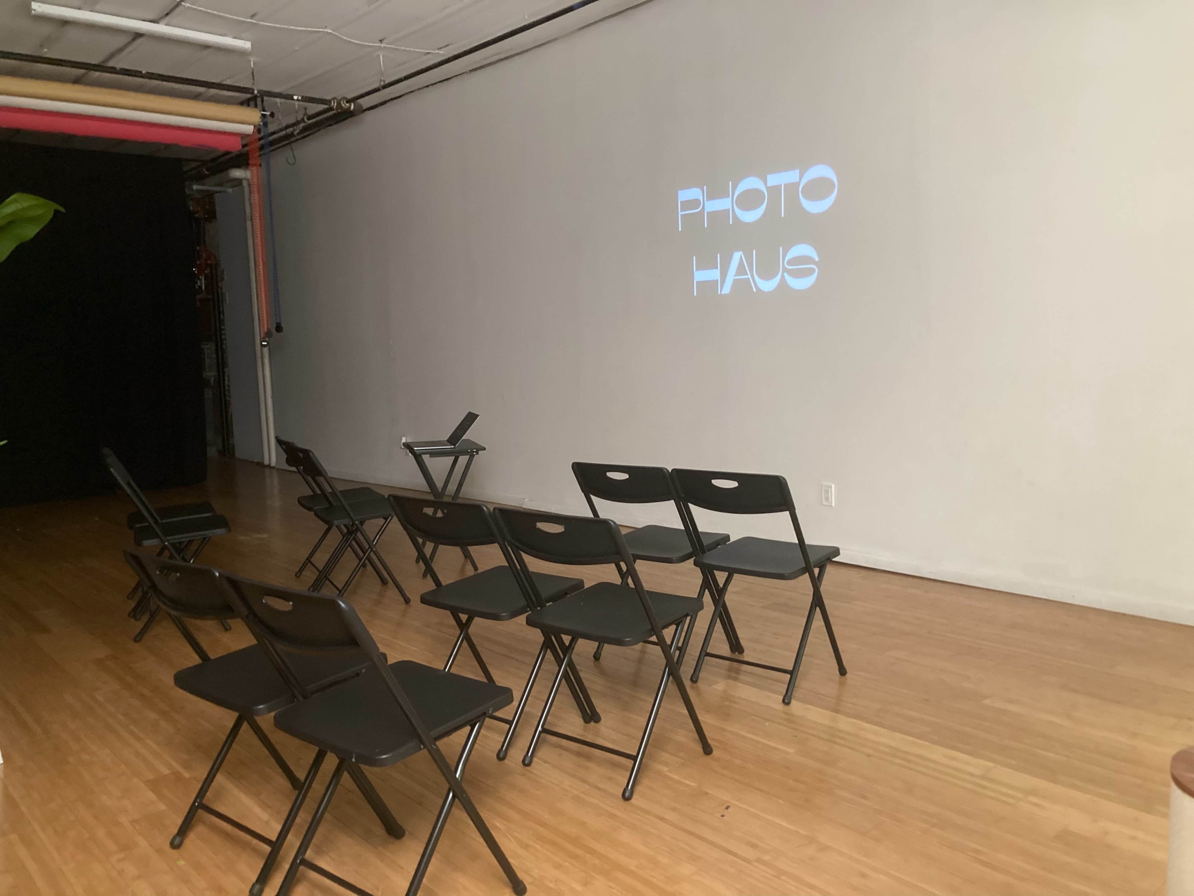 A room with wooden flooring, featuring several black folding chairs arranged in rows and a projection on the wall that reads "PHOTO HAUS."