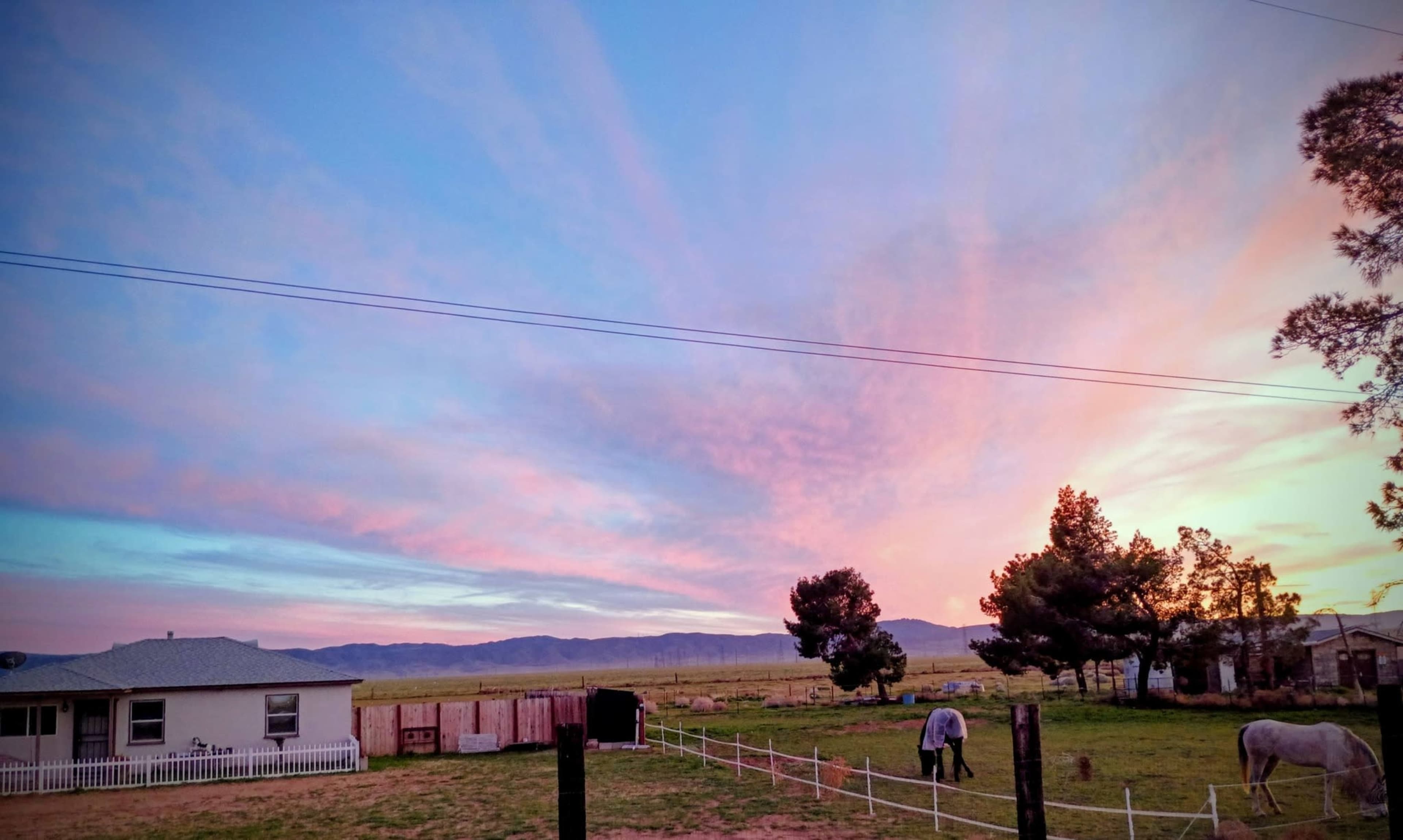 A rural landscape features a house with a front yard, surrounded by open fields and mountains under a colorful sky at sunset.