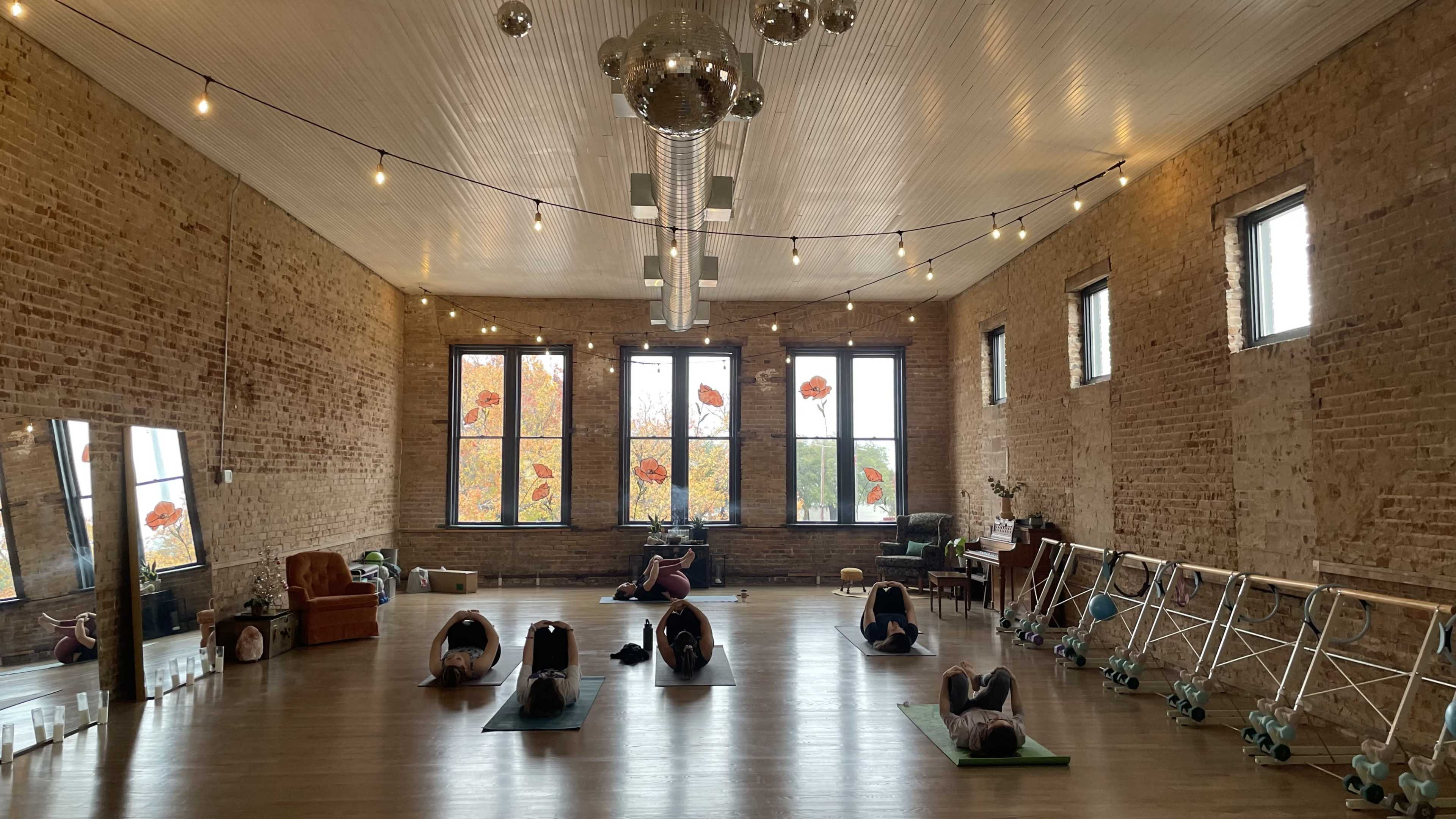 The image shows a spacious, brightly lit studio with people practicing yoga on mats, surrounded by exposed brick walls and large windows displaying autumn leaves.