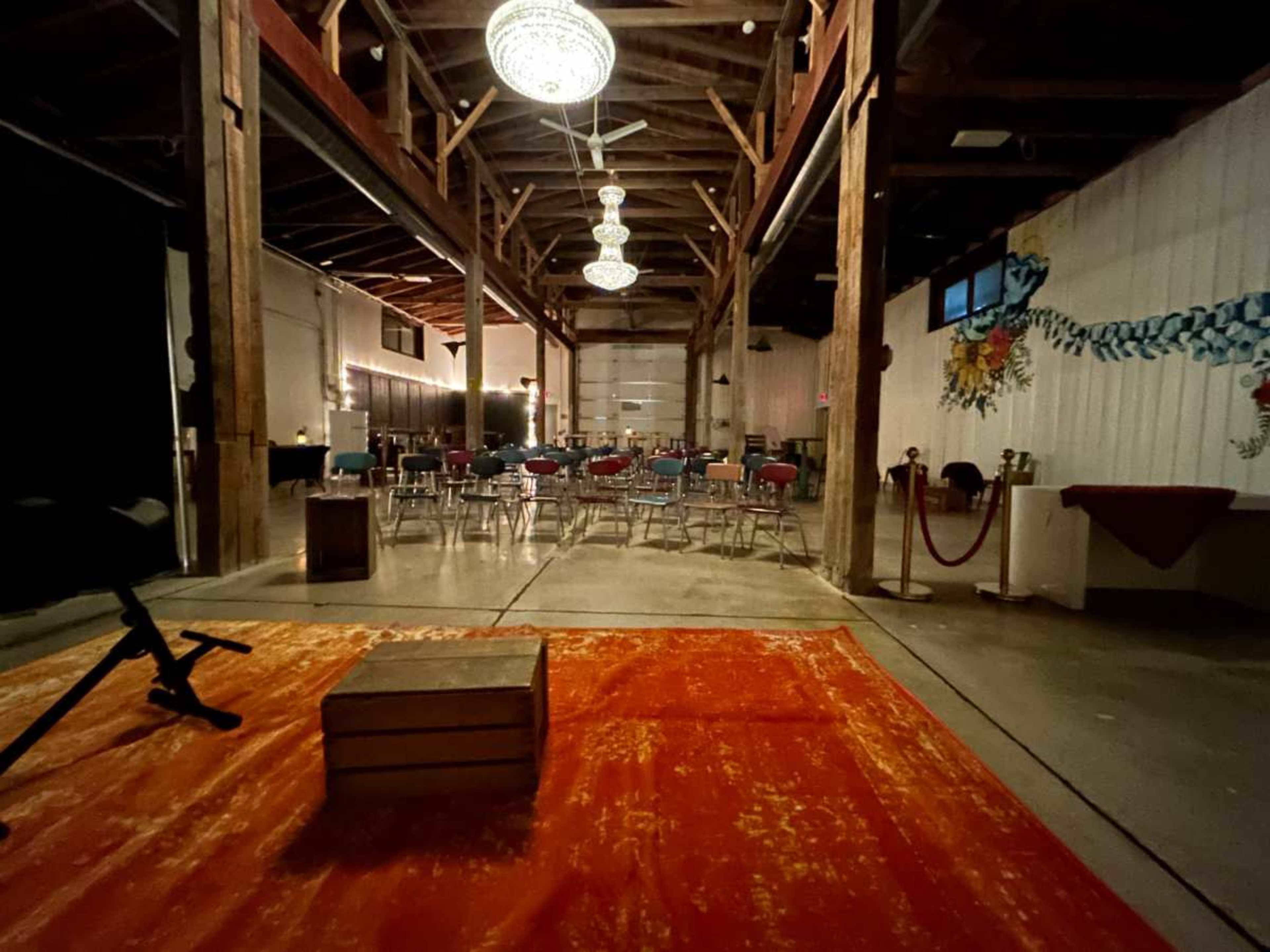 The image shows an empty event space with wooden beams, a red rug in the foreground, and rows of chairs set up in the background under hanging light fixtures.