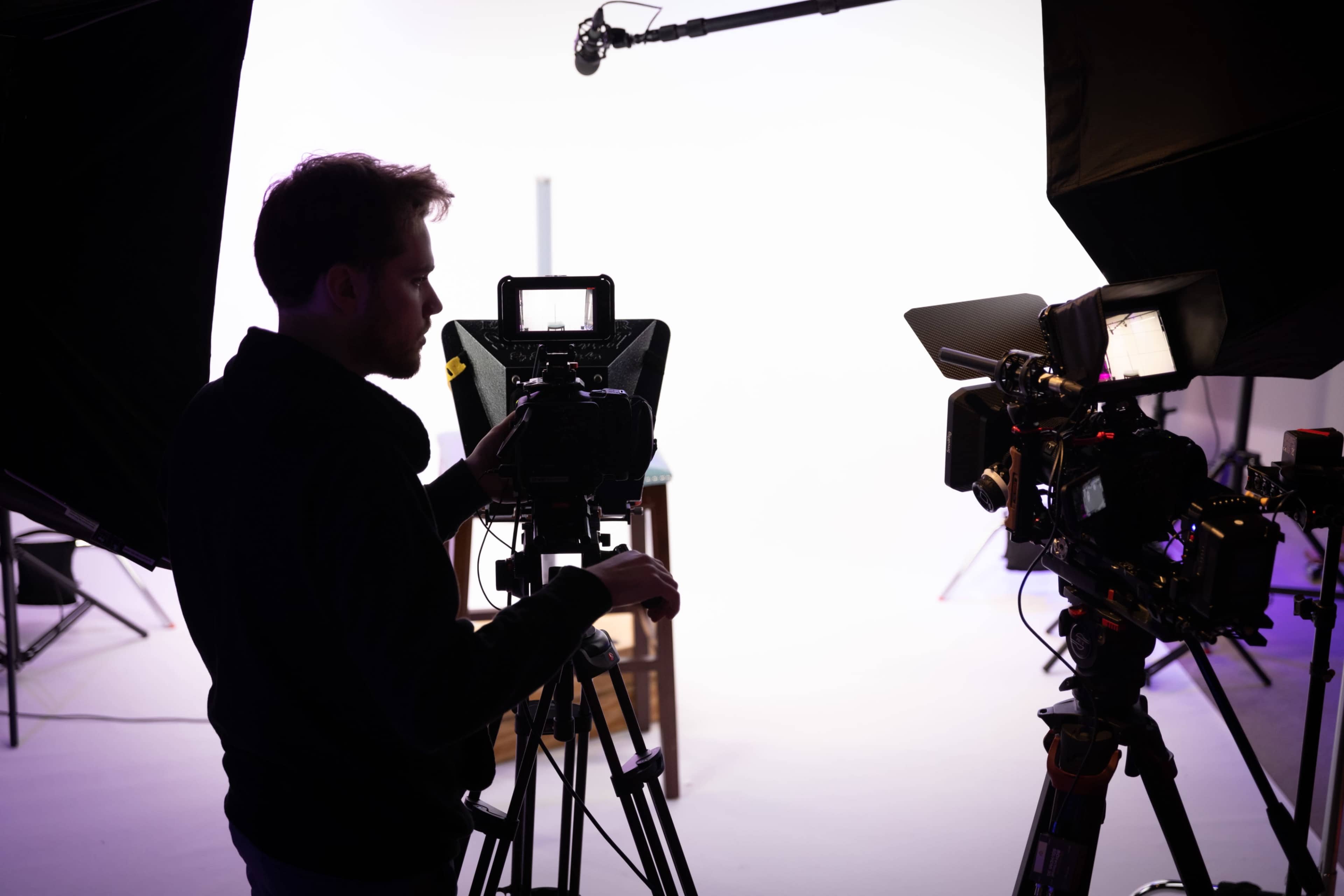 A person operates a video camera on a tripod in a studio setting with bright white lighting.