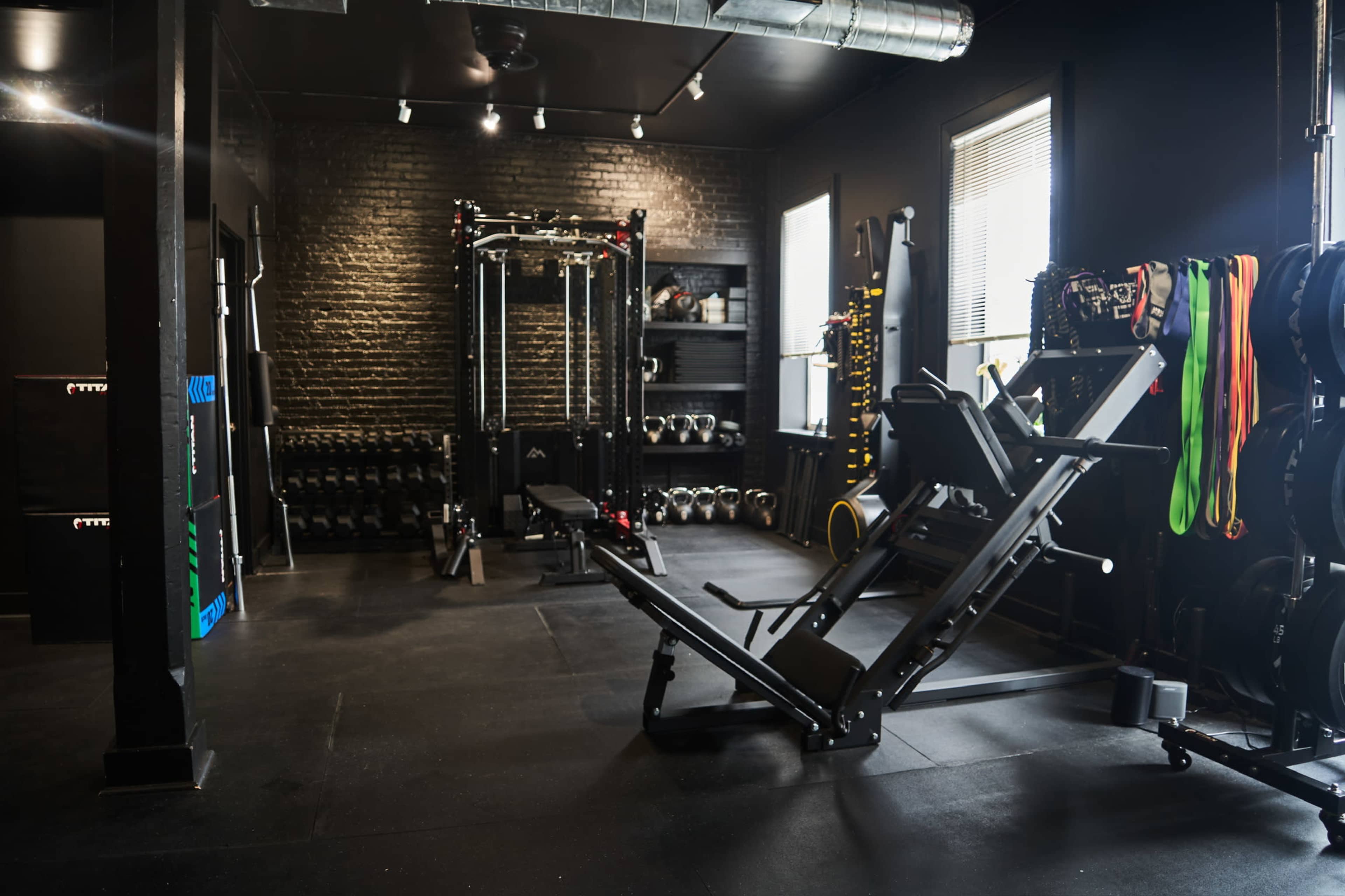The image shows a well-equipped gym space featuring weightlifting machines, dumbbells, resistance bands, and a cable machine against a black brick wall.