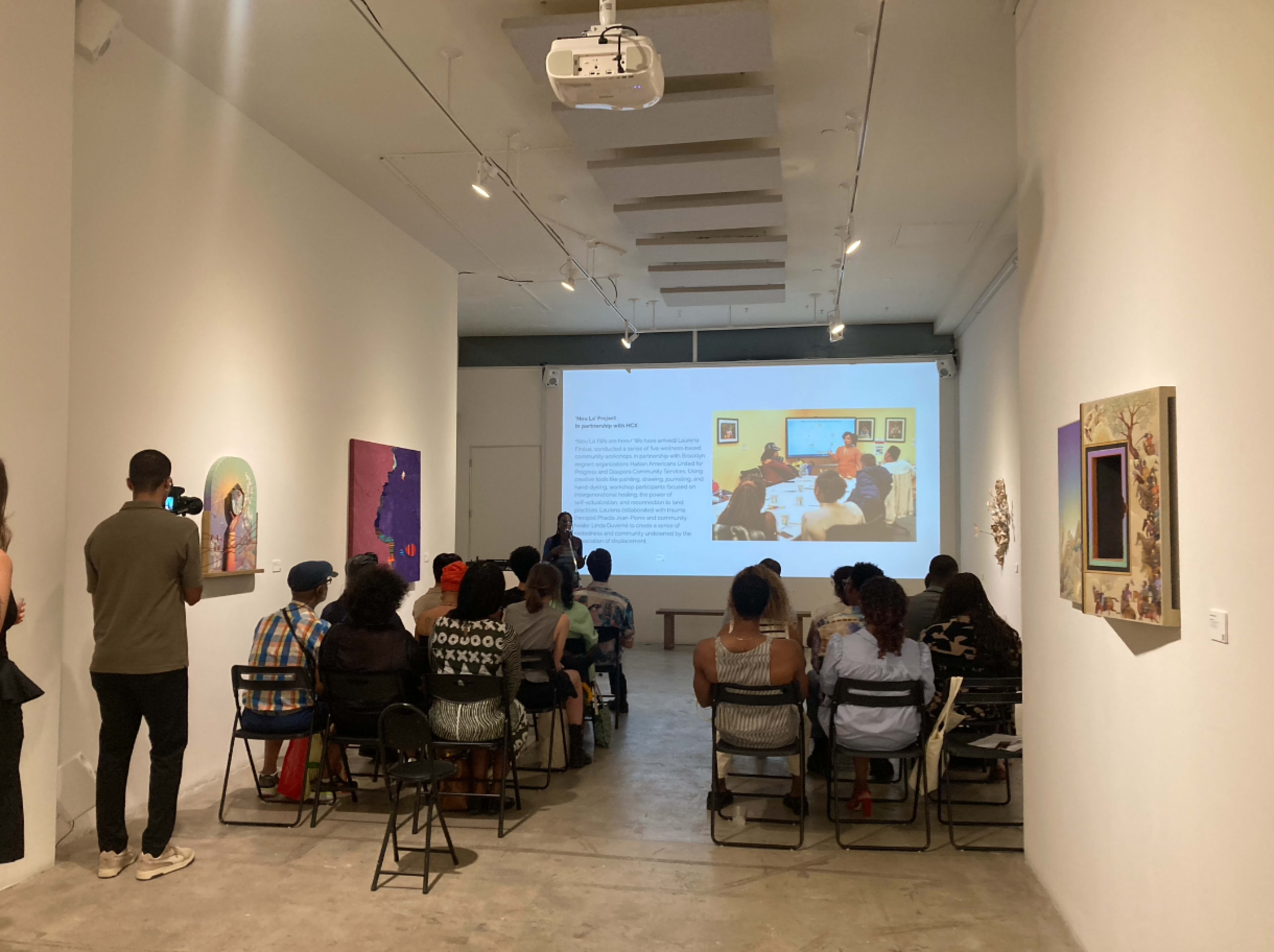 A diverse group of people sits in a gallery space, facing a projection displaying a presentation, while another individual stands nearby speaking to the audience.