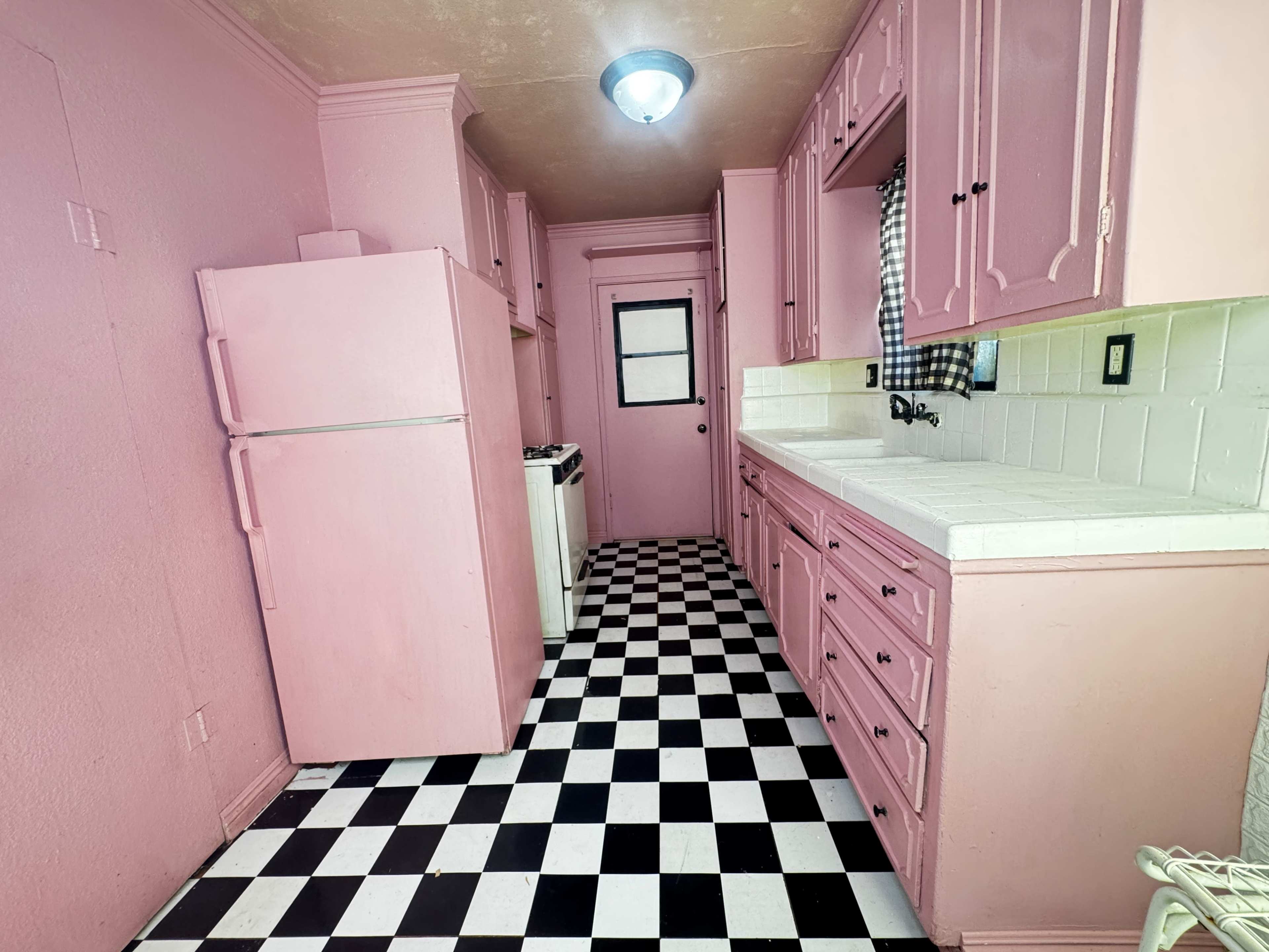 The image depicts a narrow kitchen featuring pink cabinets and walls, a black-and-white checkerboard floor, and white appliances.