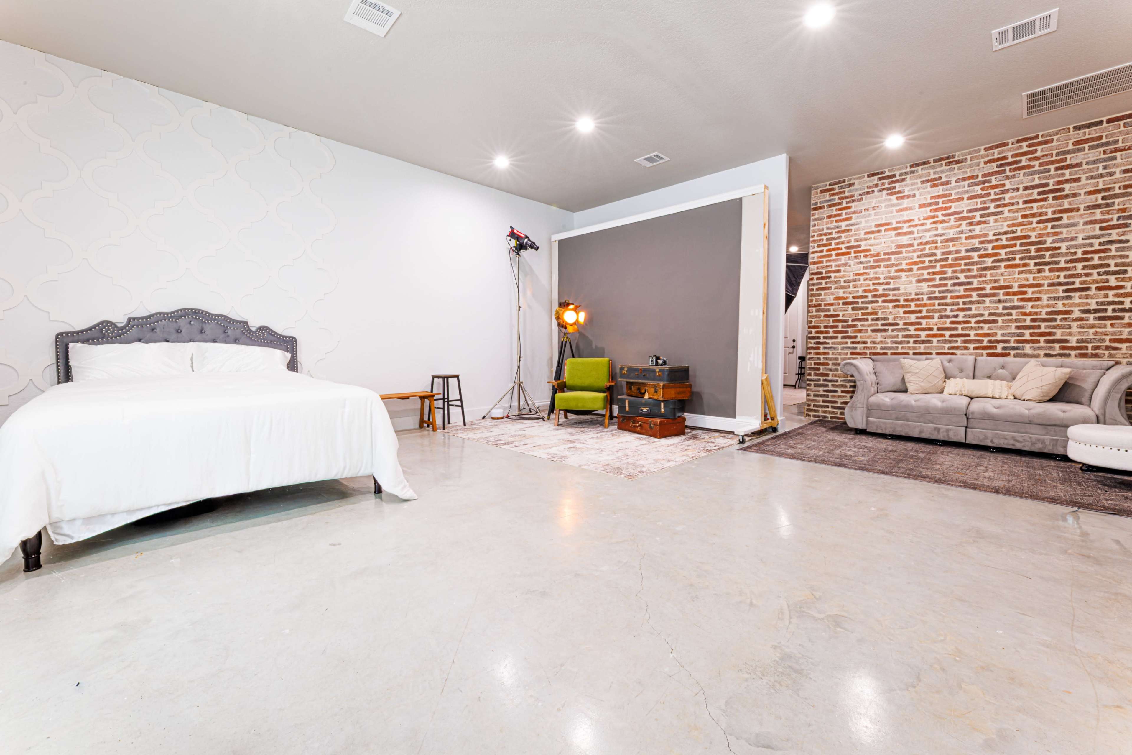 The image shows a spacious room with a bed on one side, a couch and coffee table on another, and exposed brick walls.