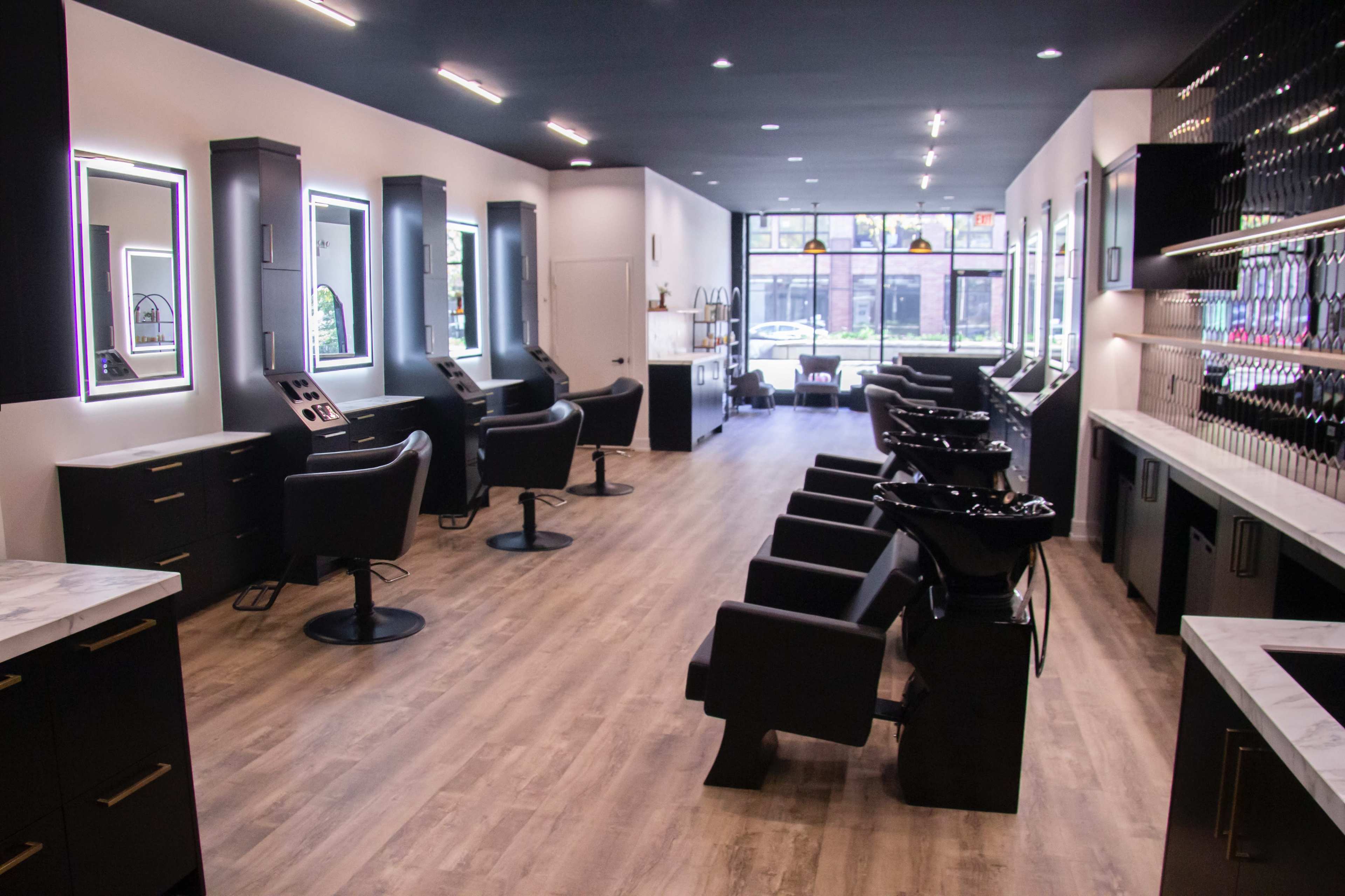 The image shows a modern hair salon with several styling stations and wash basins, featuring sleek black furniture and bright lighting.