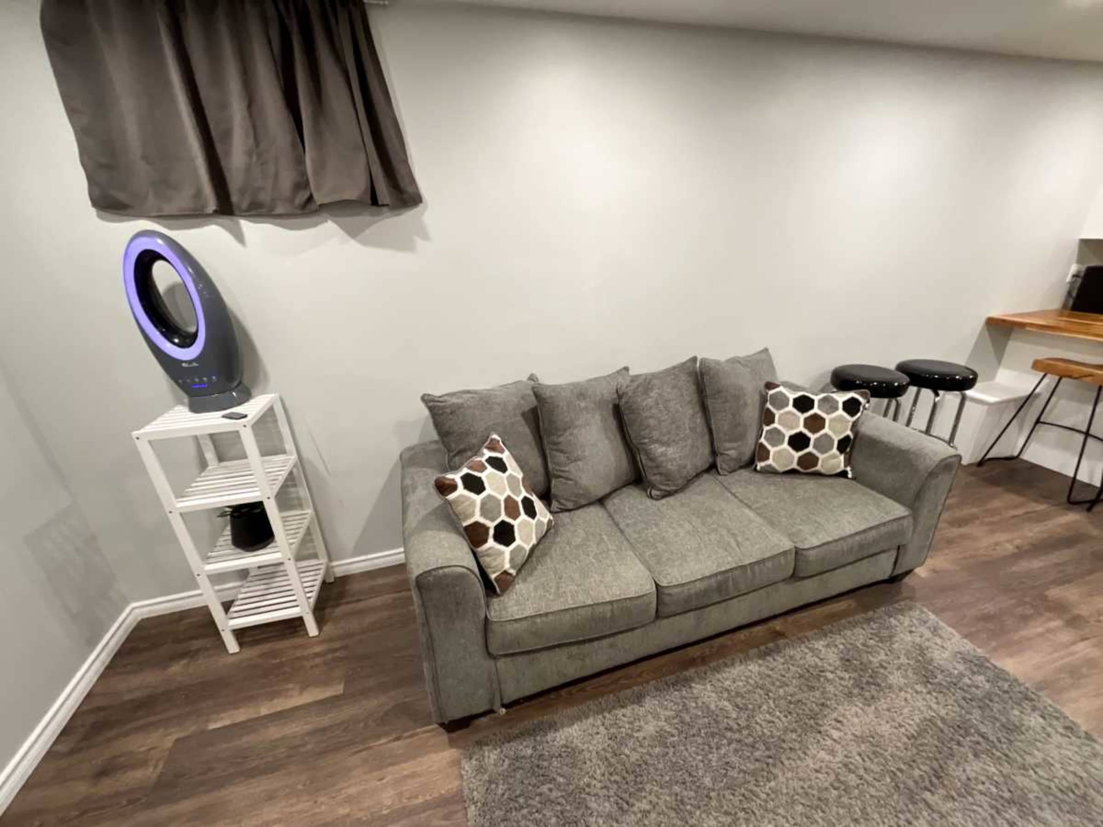The image shows a gray sofa with polka dot pillows, a white shelving unit, a modern fan, a bar area with stools, and a light-colored wall in a basement room.