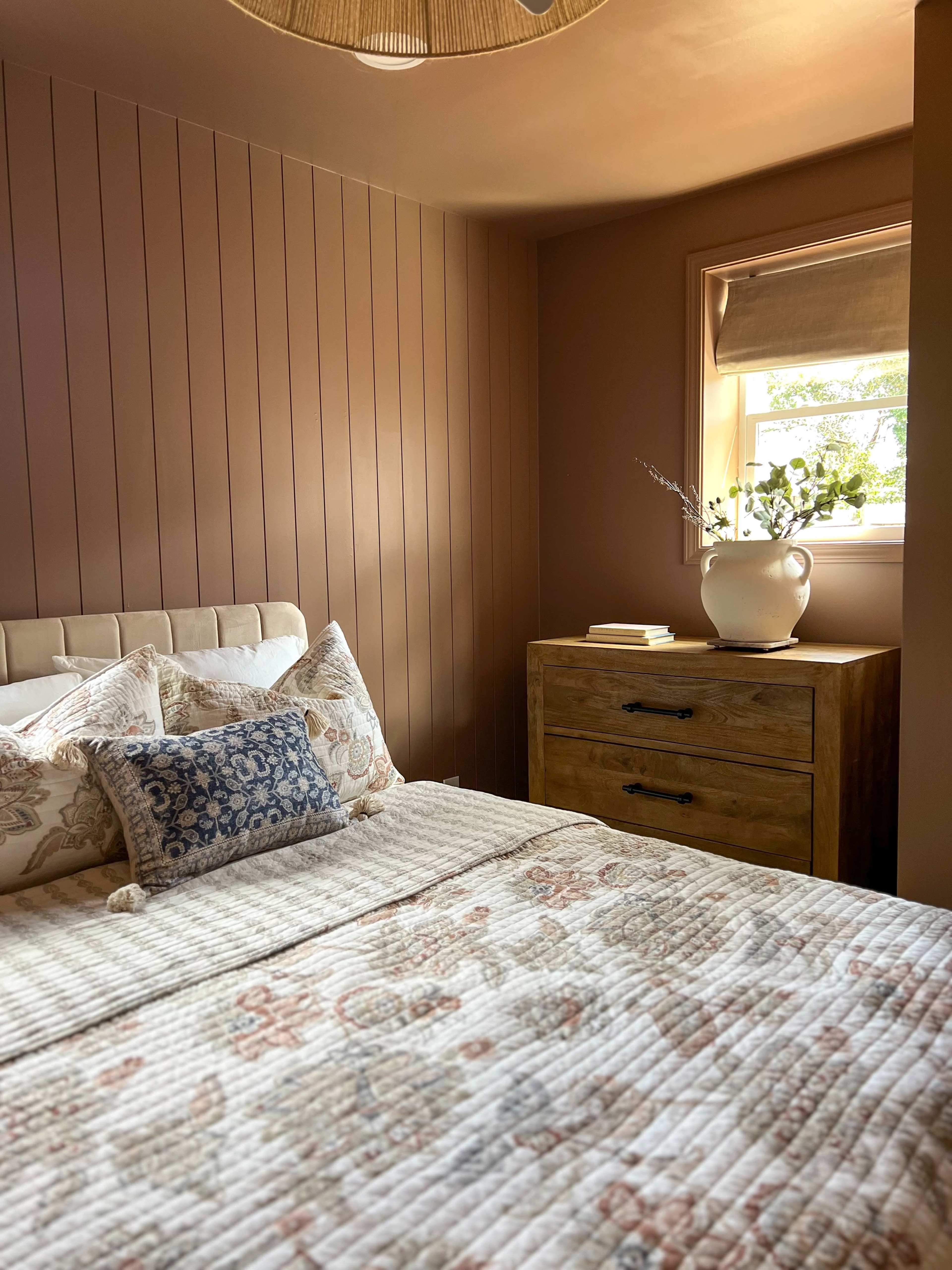 A cozy bedroom with a bed adorned with decorative pillows and a wooden dresser beside a window.