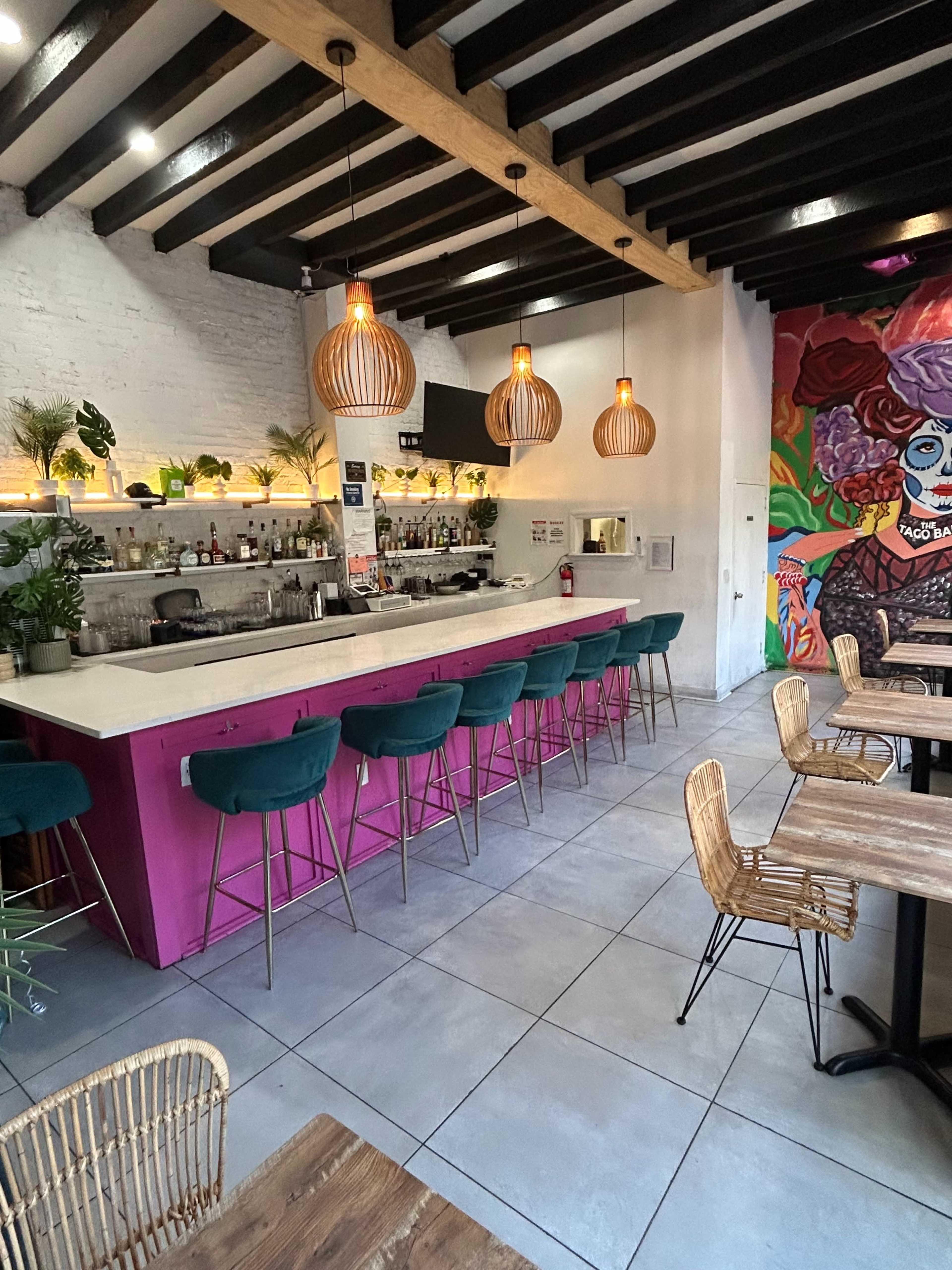 The image shows the interior of a stylish restaurant with a bar area, featuring colorful seating and a vibrant mural on one wall.