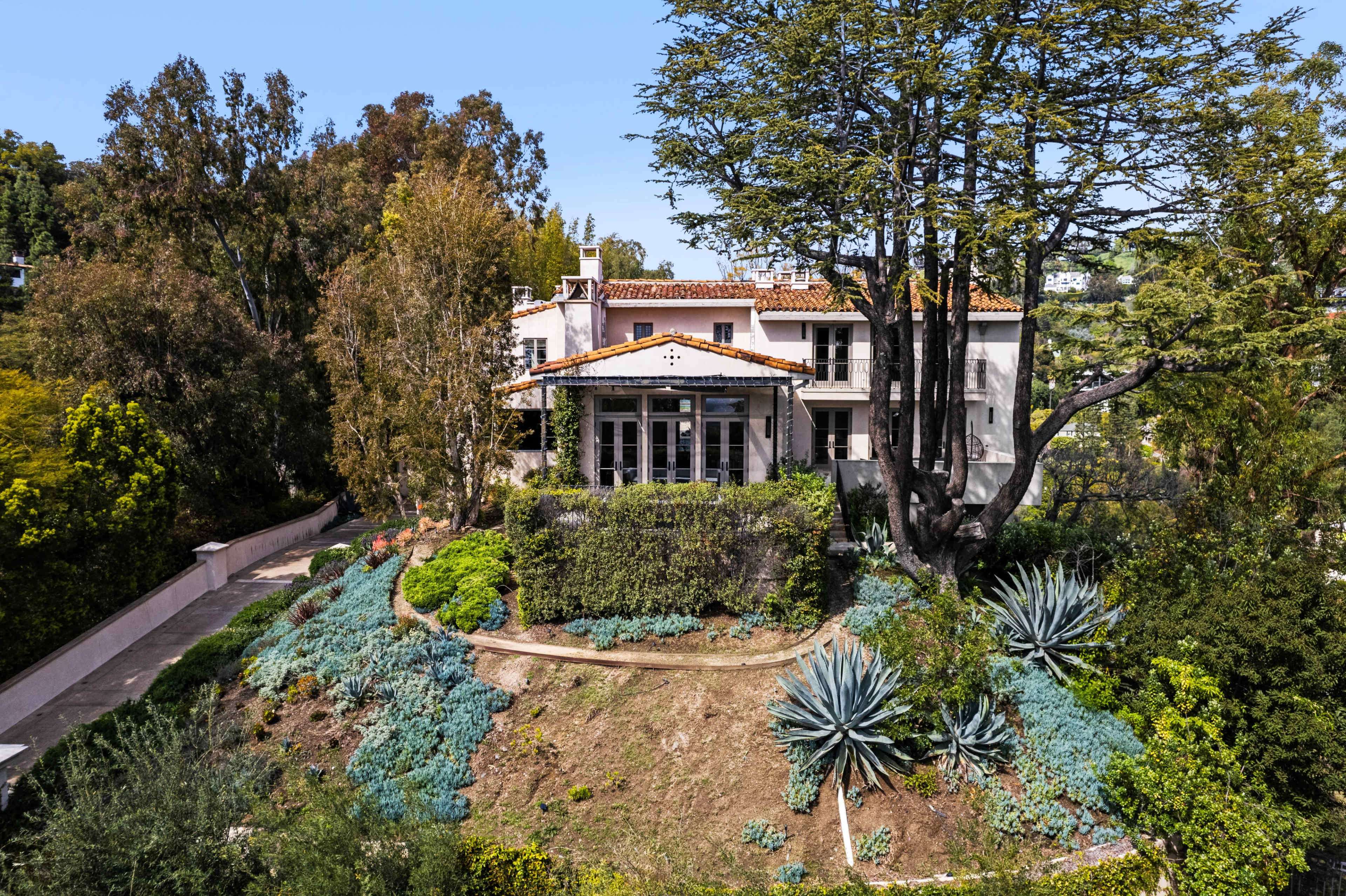 A large house is situated on a hillside surrounded by various trees and plants, with a well-maintained garden in the foreground.