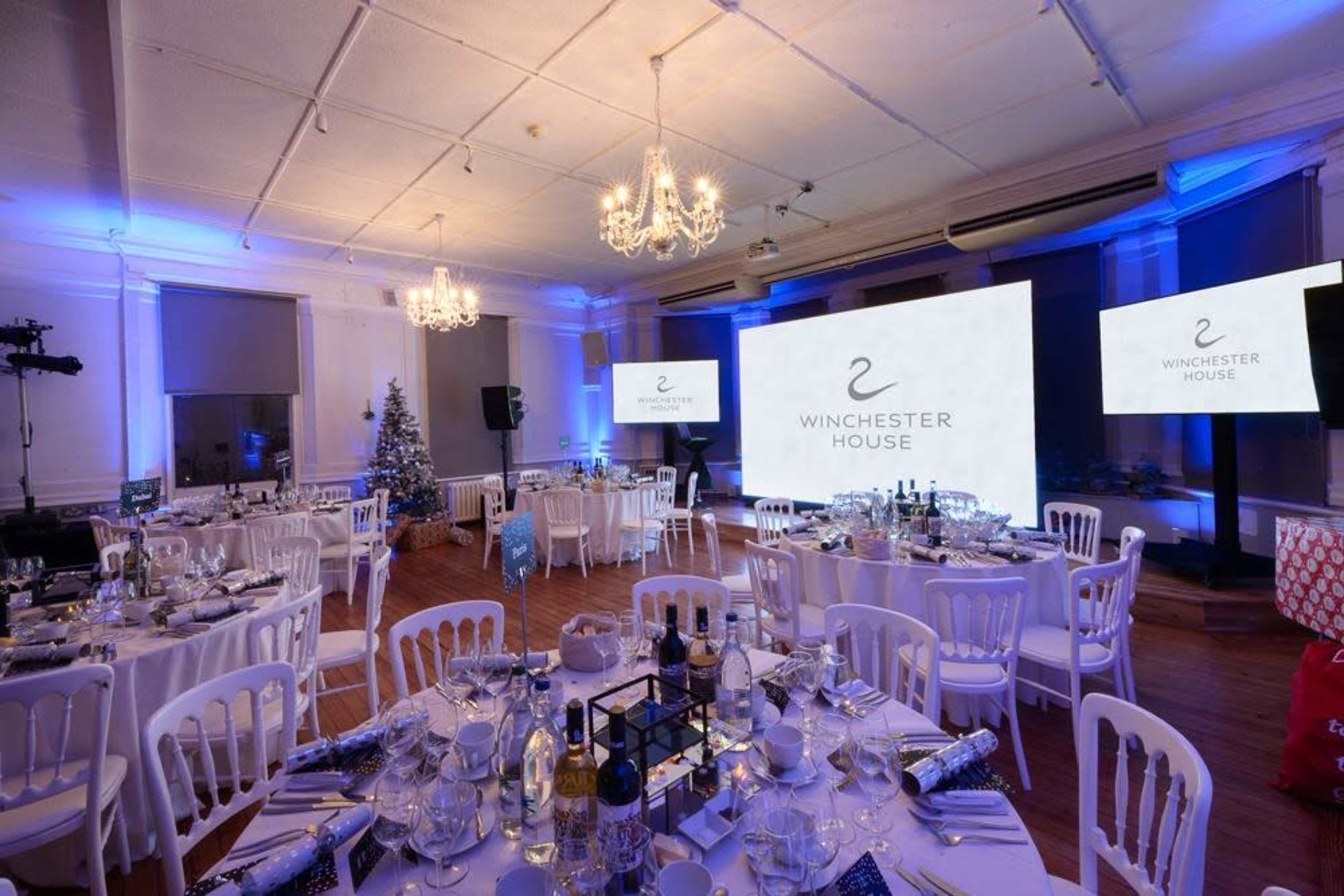A formally set banquet hall with tables arranged for an event, featuring blue lighting and large screens displaying the "Winchester House" logo.