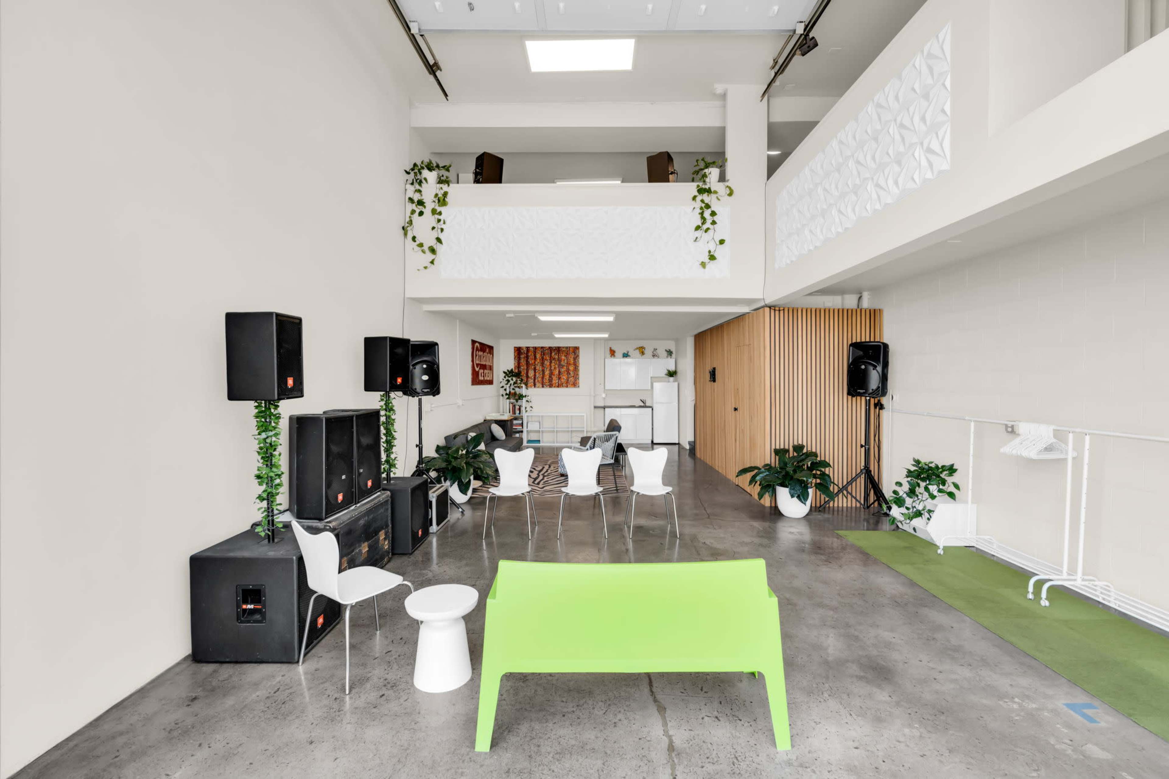 The image shows a spacious indoor area with a green seating arrangement, white chairs, sound equipment, and plants, featuring high ceilings and an upper balcony.