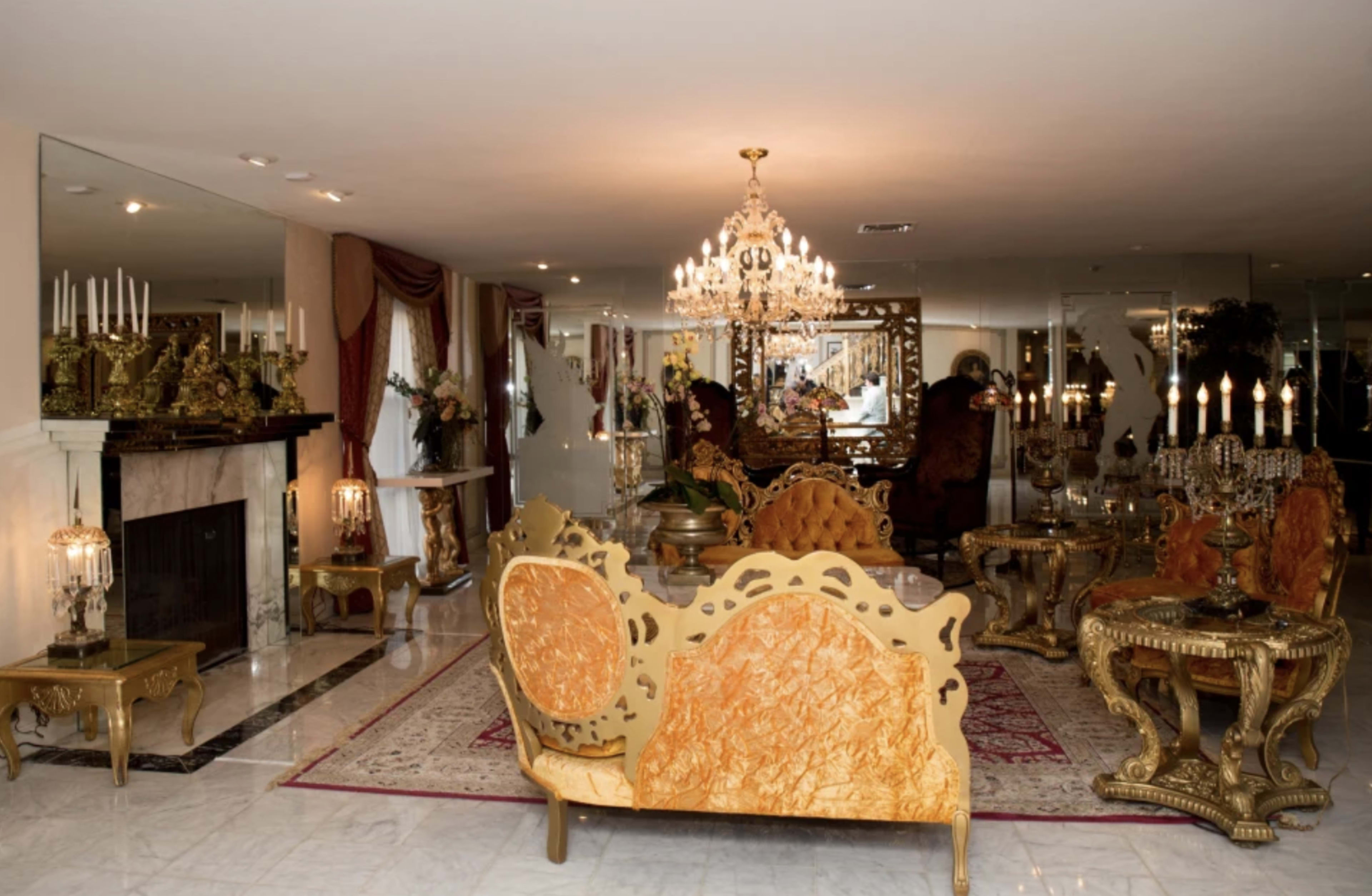 The room features an ornate gold and marble decor with a chandelier, a large mirror, and elegant furniture arranged around a central coffee table.