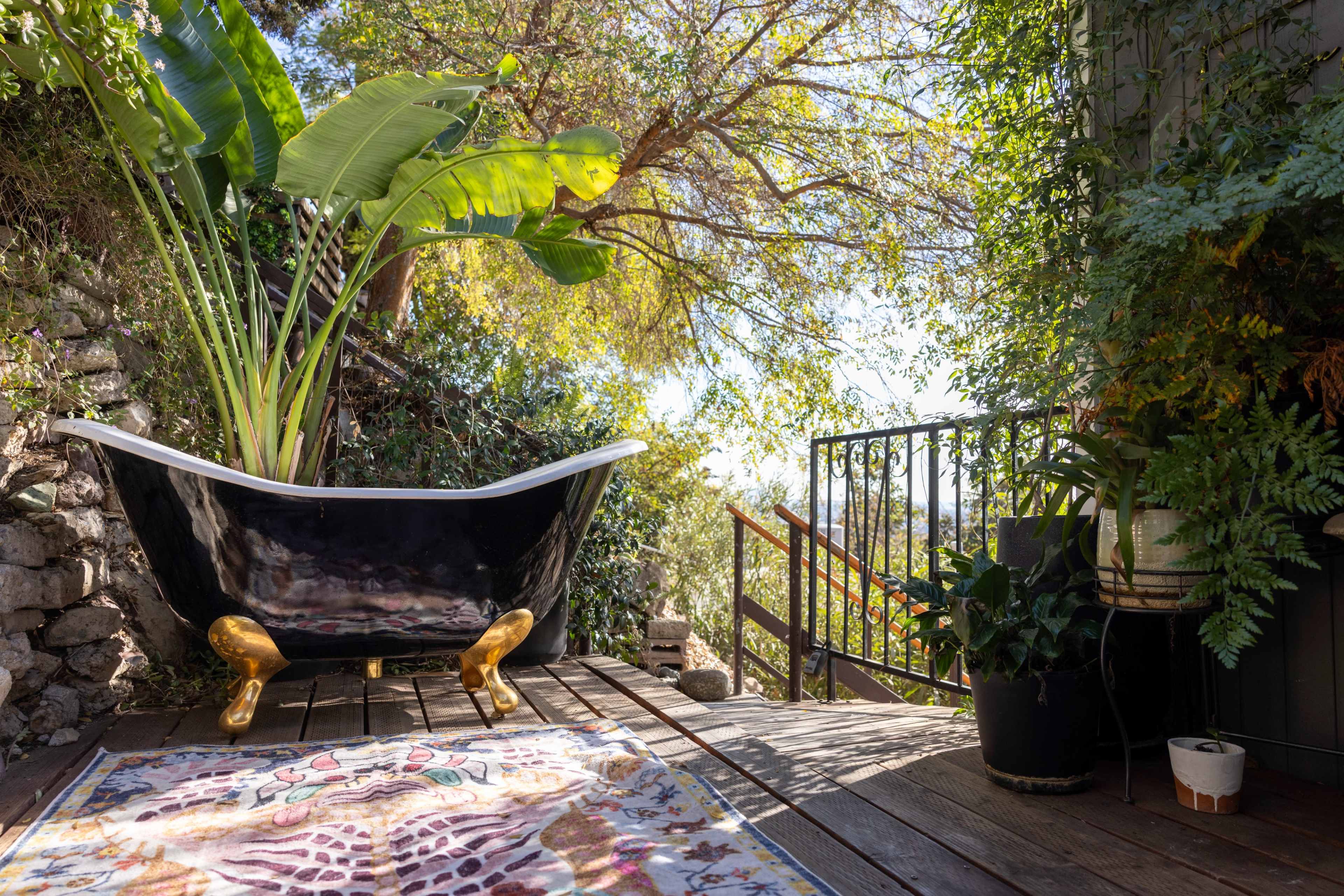 A vintage black clawfoot bathtub with golden legs is adorned with large green plants, placed on a wooden deck surrounded by trees and decorative potted plants.