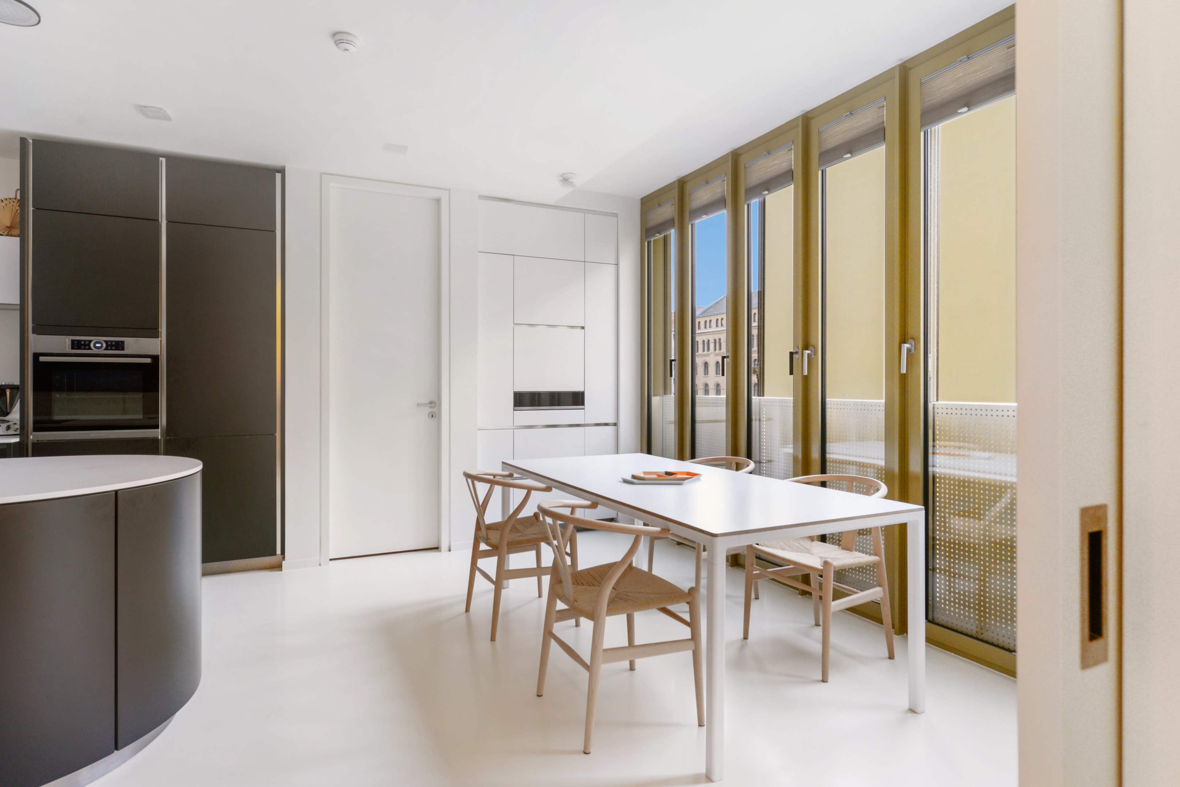 A modern kitchen features a white table surrounded by wooden chairs, with large windows allowing natural light to enter.