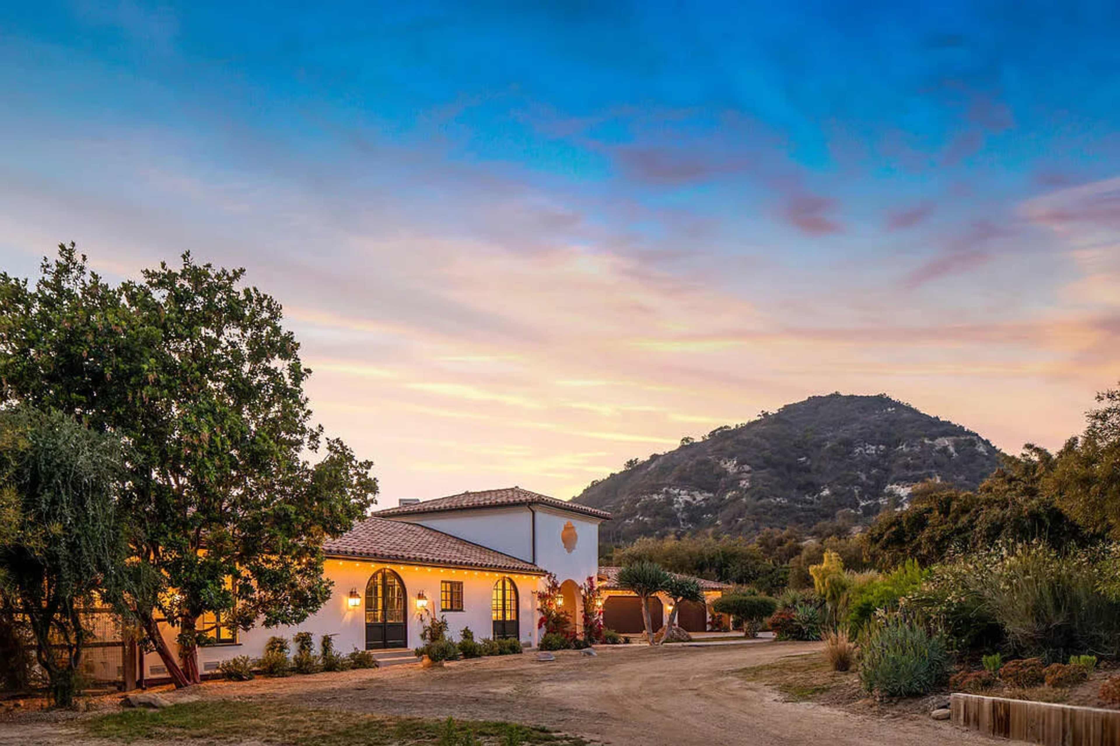 A white stucco house with a tiled roof sits at the base of a hill, surrounded by greenery under a colorful sky at dusk.