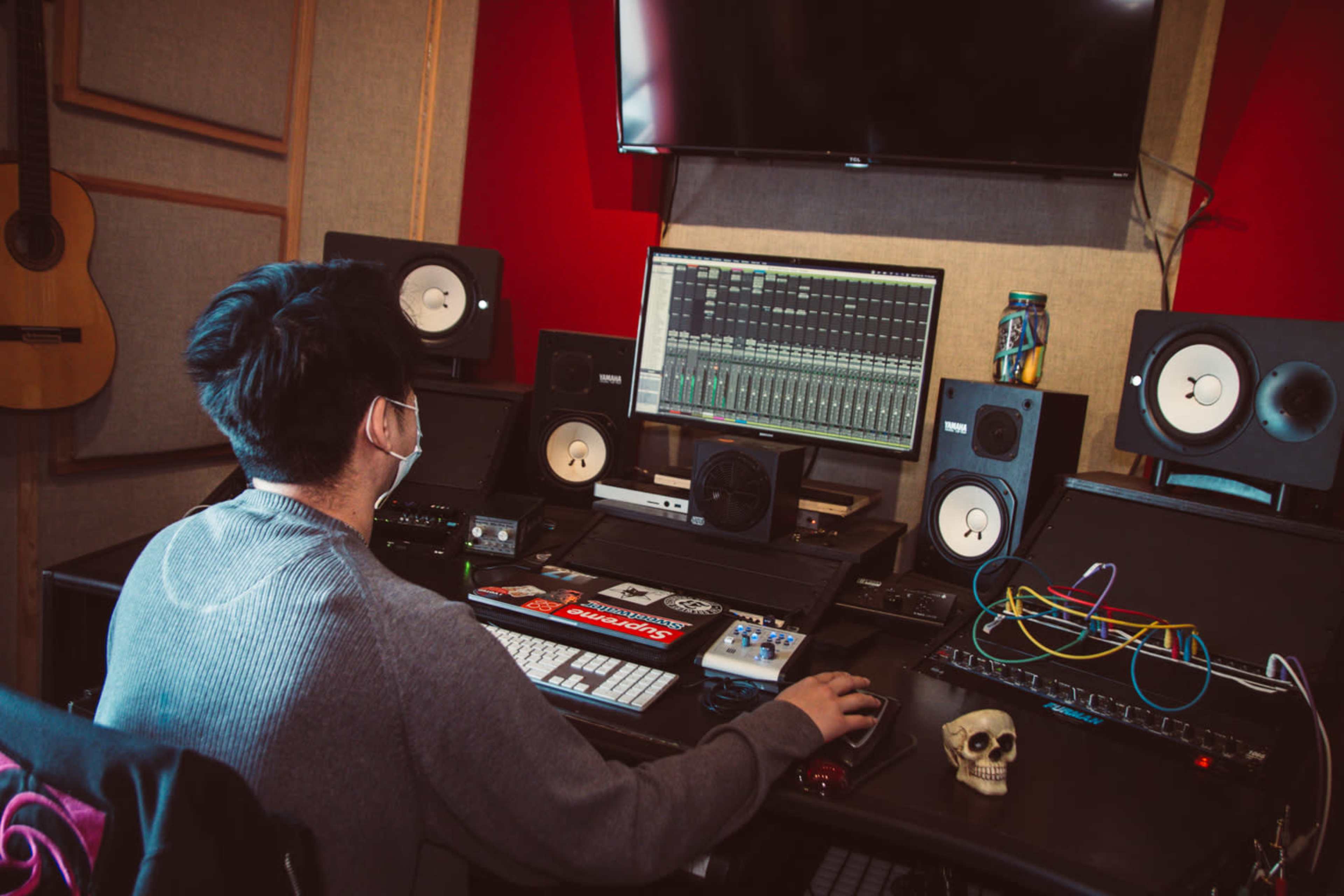 A person wearing a mask sits at a sound mixing desk surrounded by audio equipment and speakers, with a computer screen displaying audio tracks.