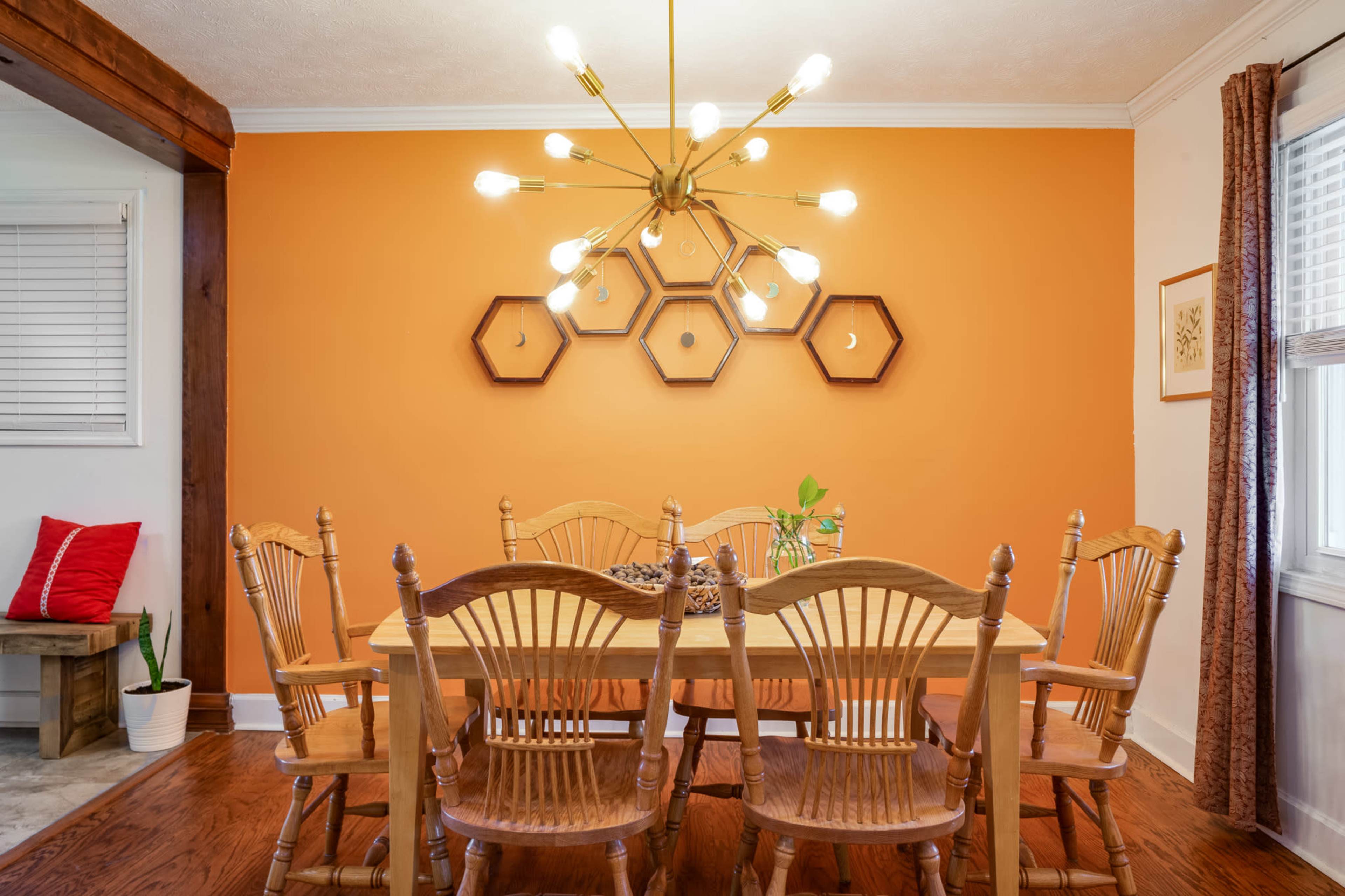 A dining room features a wooden table surrounded by six chairs, with a striking orange wall adorned by hexagonal wooden frames and a modern light fixture above.