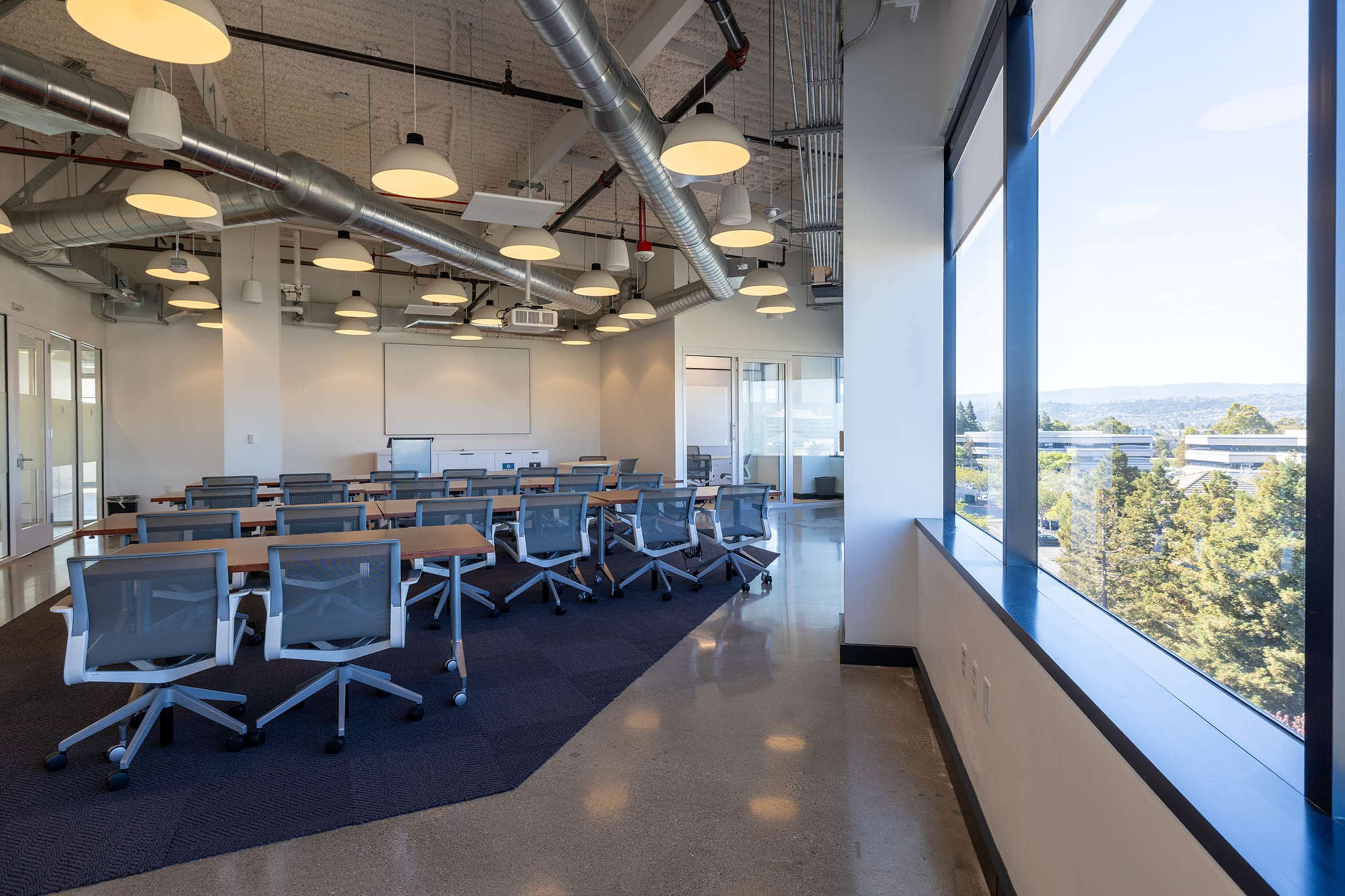 The image shows a modern classroom with rows of desks and chairs, large windows, and bright overhead lighting.