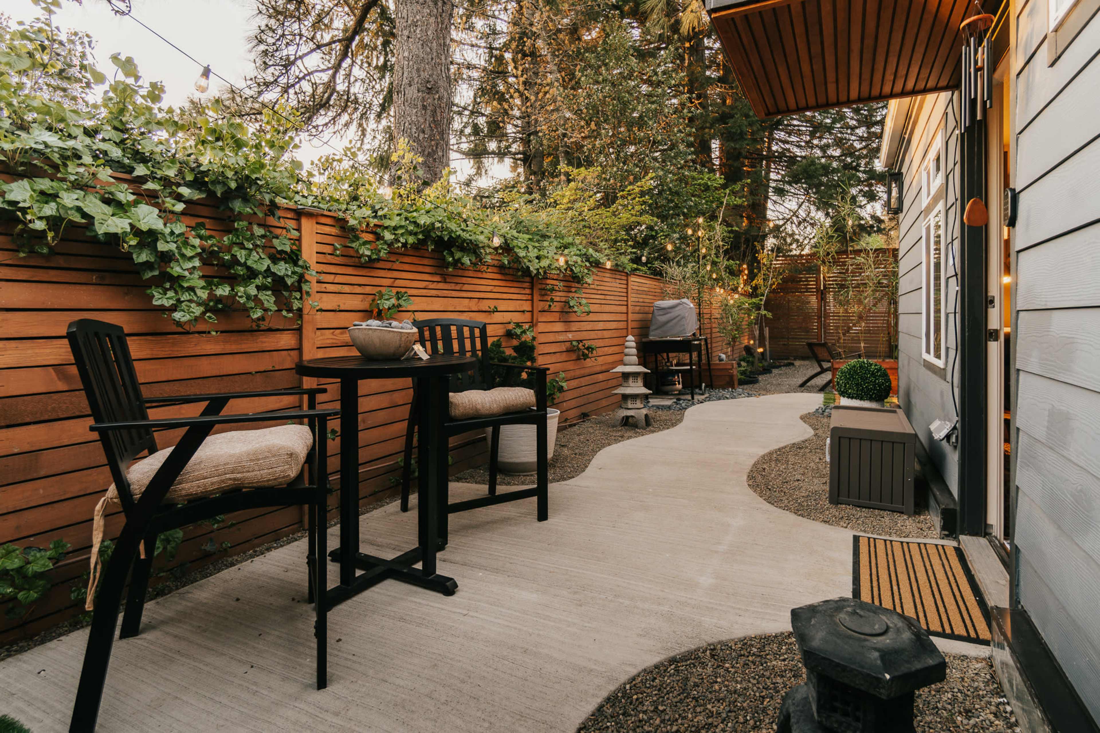 The image shows a narrow outdoor path with a concrete surface, flanked by wooden fences covered in greenery, leading to a cozy seating area with two chairs and a small table.