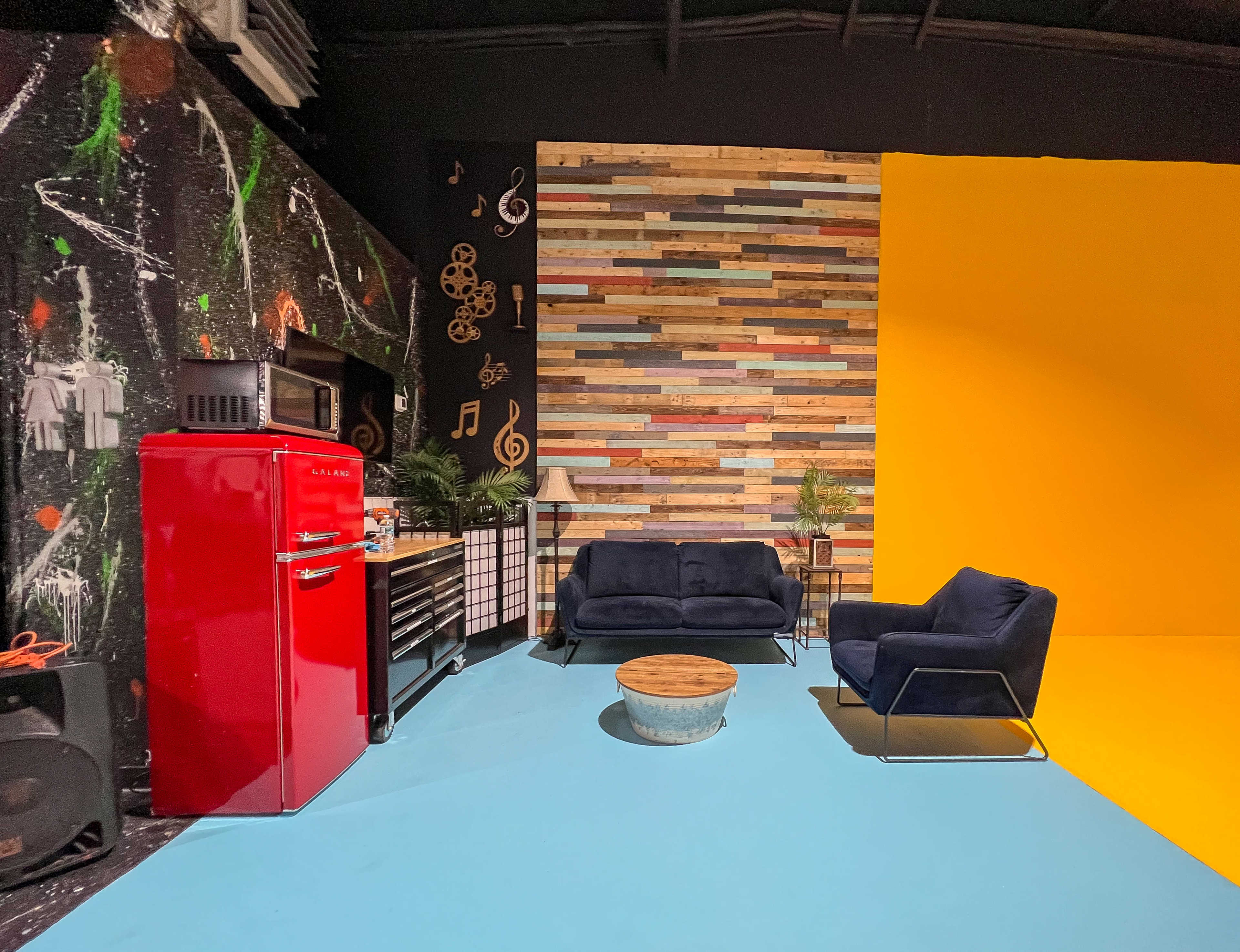 The image depicts a vibrant studio space featuring a red refrigerator, colorful wooden accent wall, and two dark sofas arranged around a round coffee table on a blue floor.