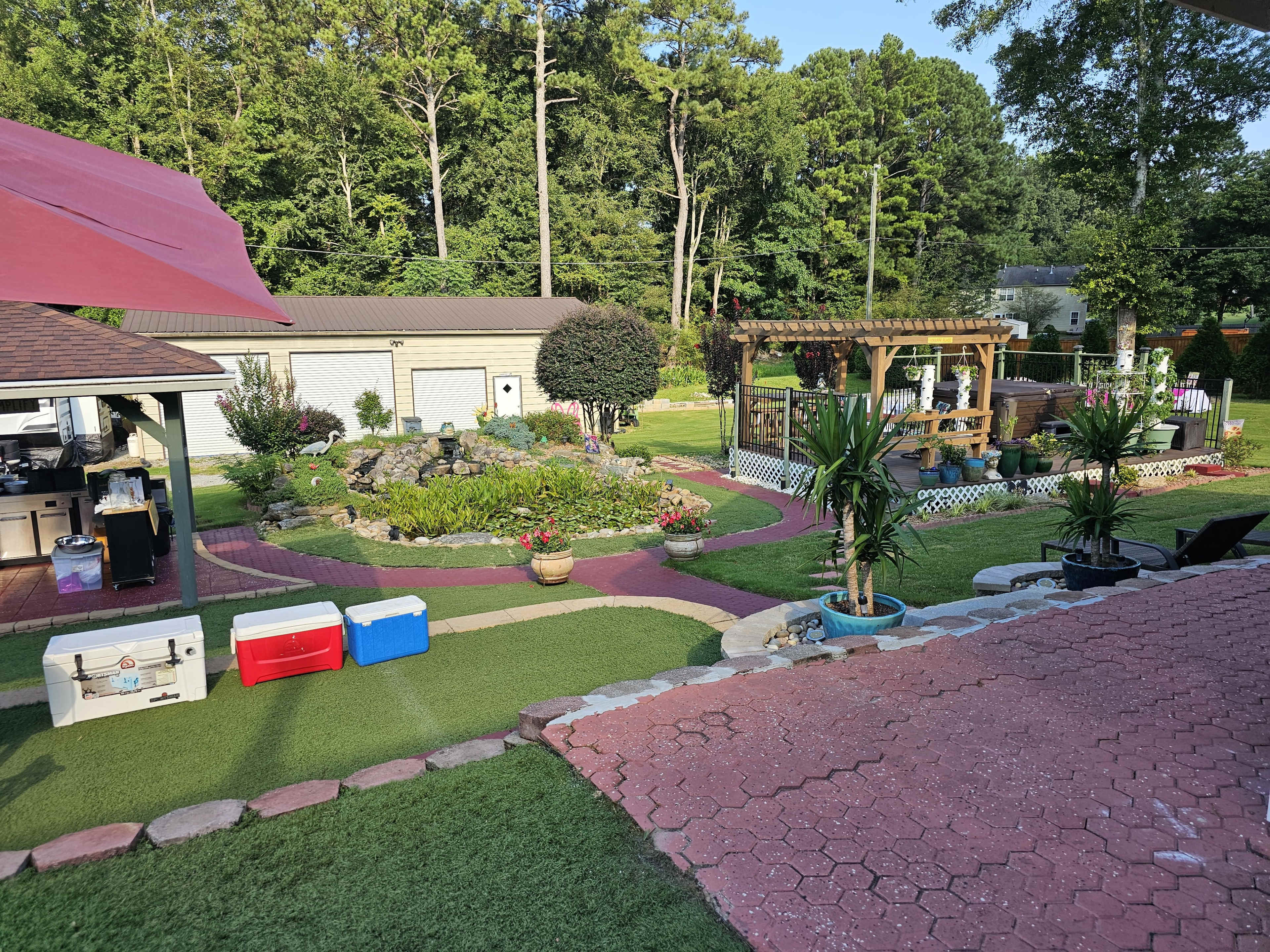 The image shows a landscaped backyard with a patio area, flower beds, garden structures, and a small water feature surrounded by greenery.