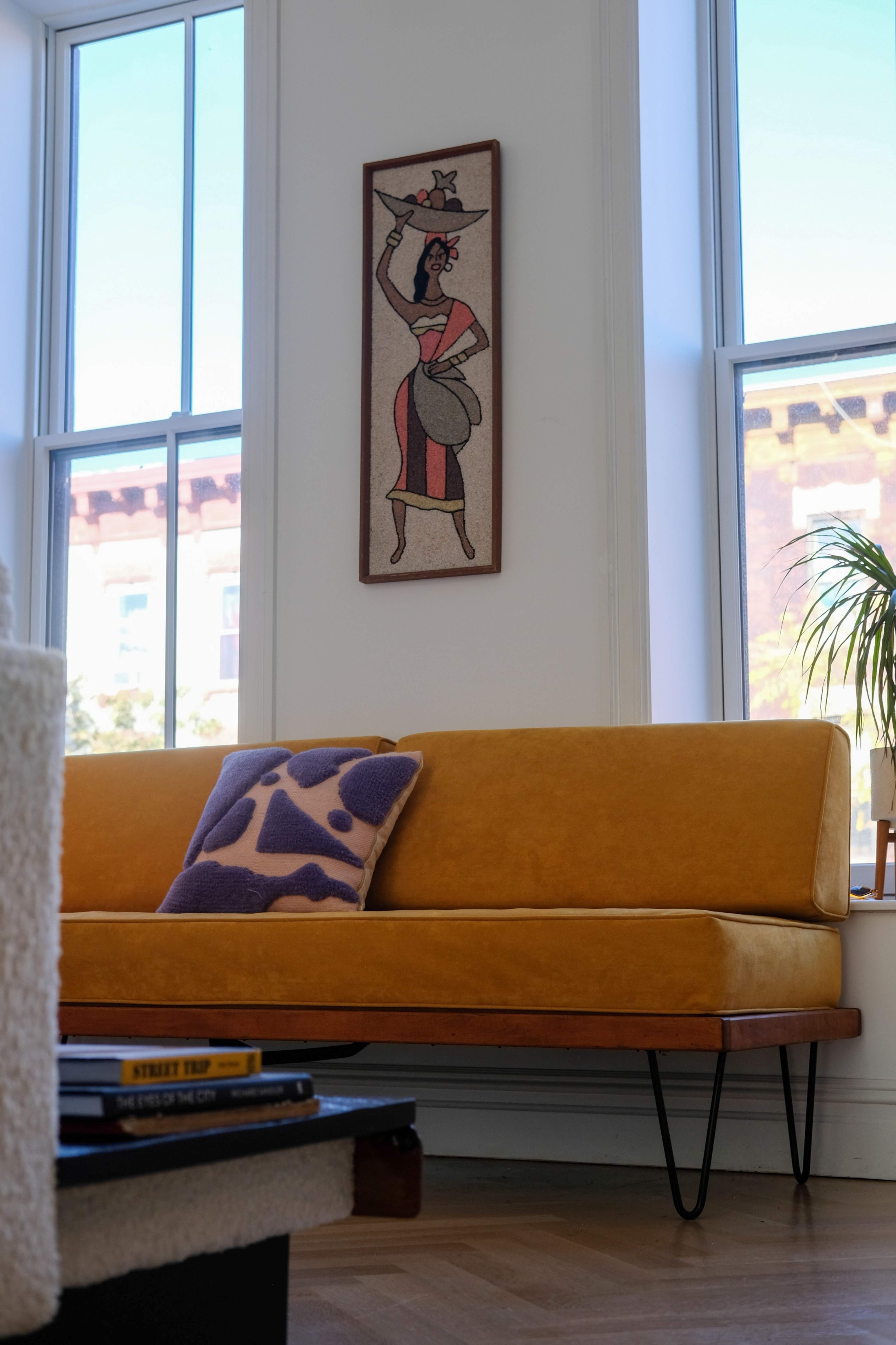 The image shows a yellow couch with a decorative pillow, positioned near large windows and decorated with a wall hanging of a woman.