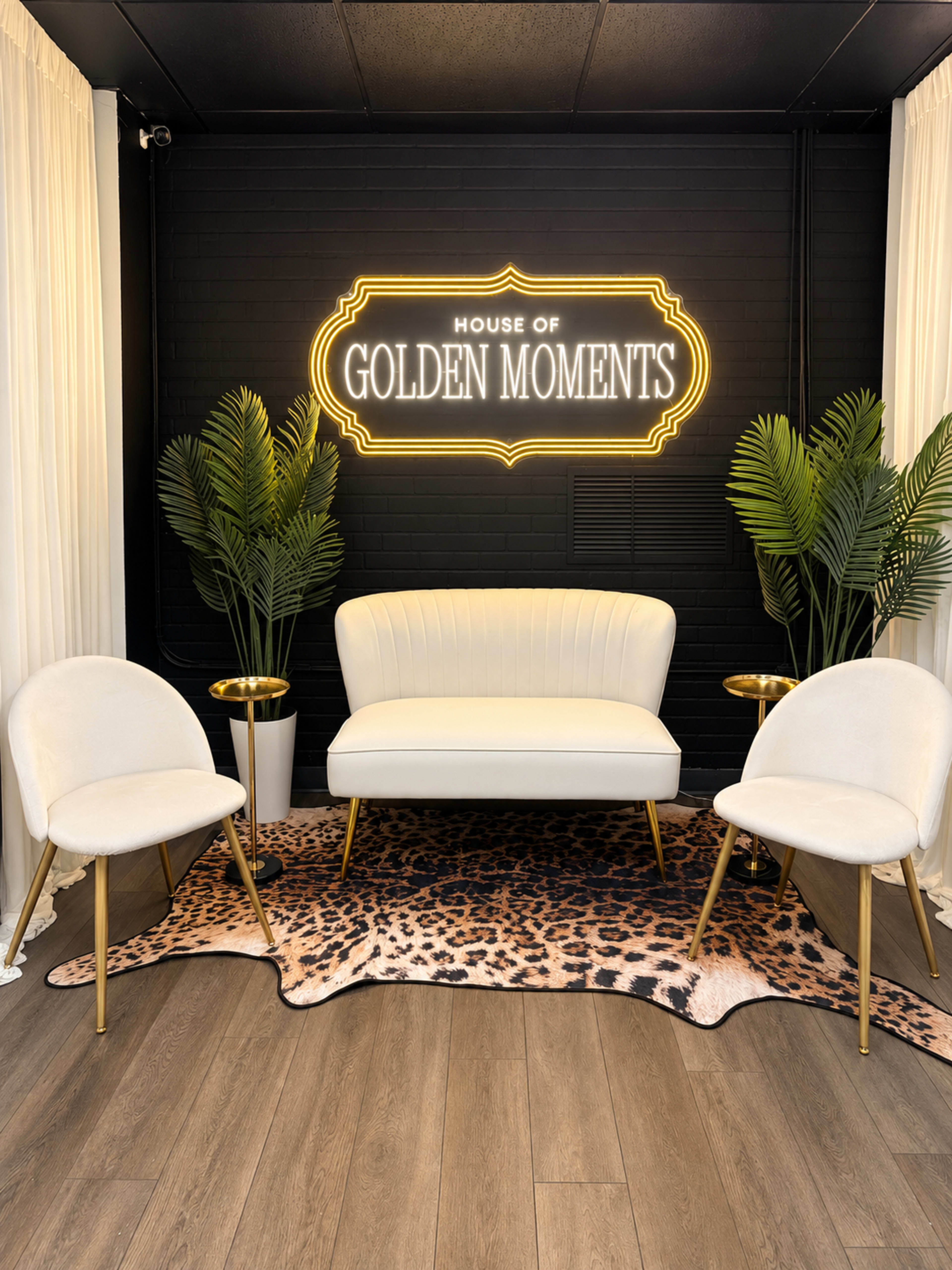 The image shows a well-decorated seating area featuring a white loveseat, two white chairs, a leopard print rug, and potted plants, all under a illuminated sign that reads "HOUSE OF GOLDEN MOMENTS."