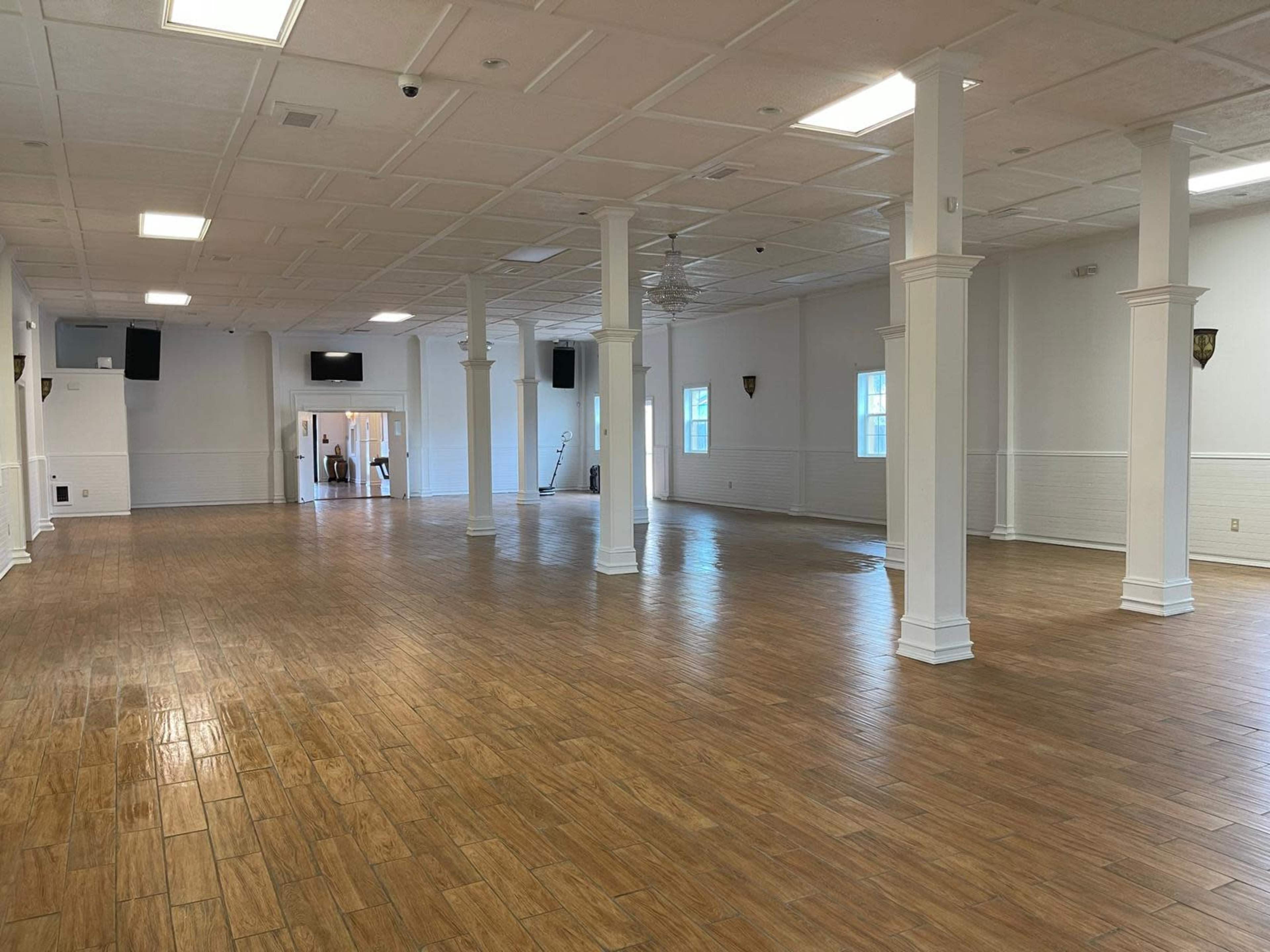 The image shows an empty, spacious room with wooden flooring, white walls, and tall columns, illuminated by overhead lights.