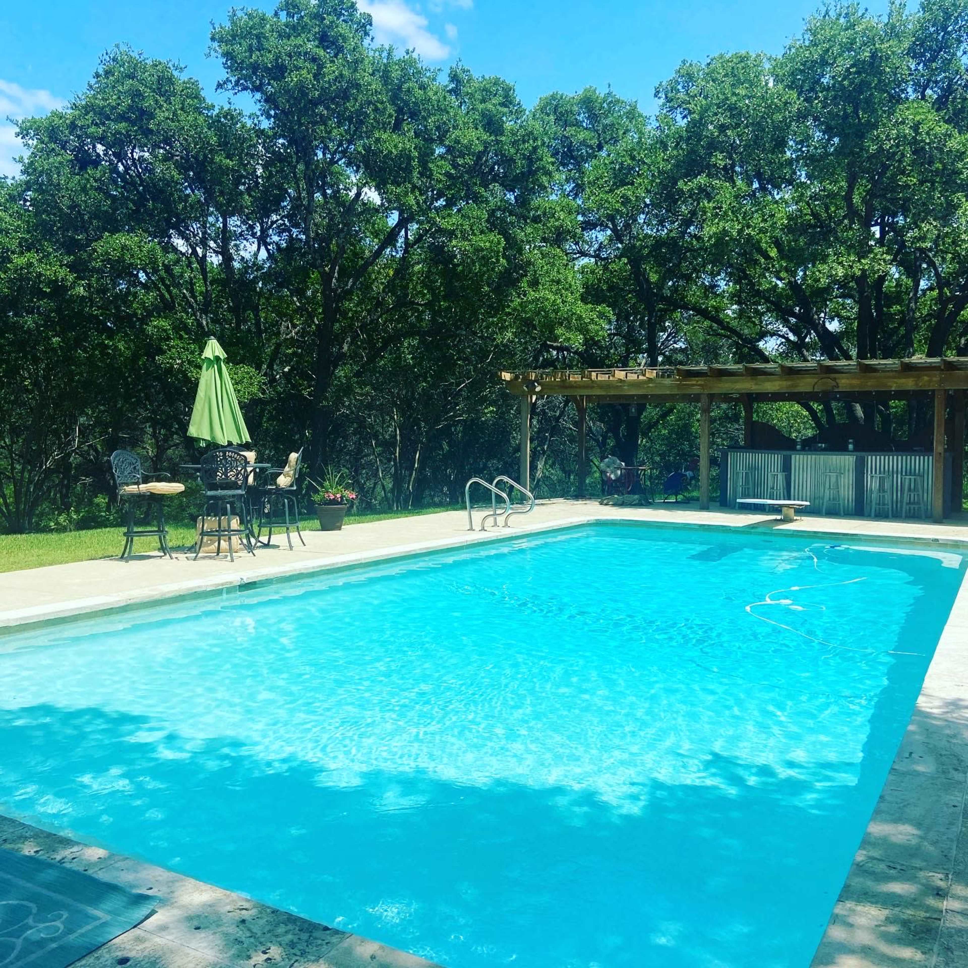 A clear swimming pool is surrounded by greenery and features a shaded seating area with an umbrella.