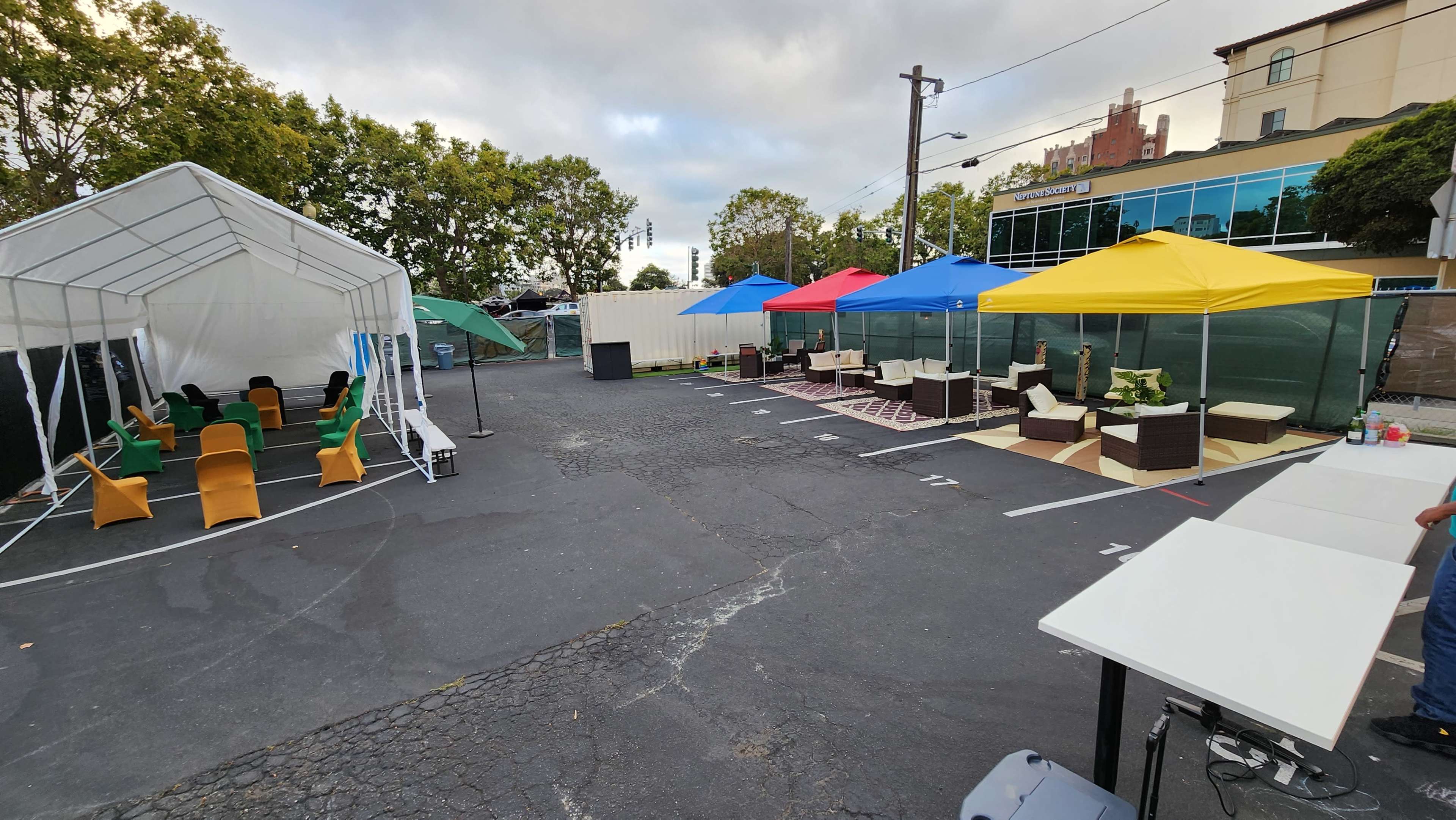 An outdoor event setup in a parking lot, featuring canopies with colorful roofs, seating arrangements, and partitioned areas.