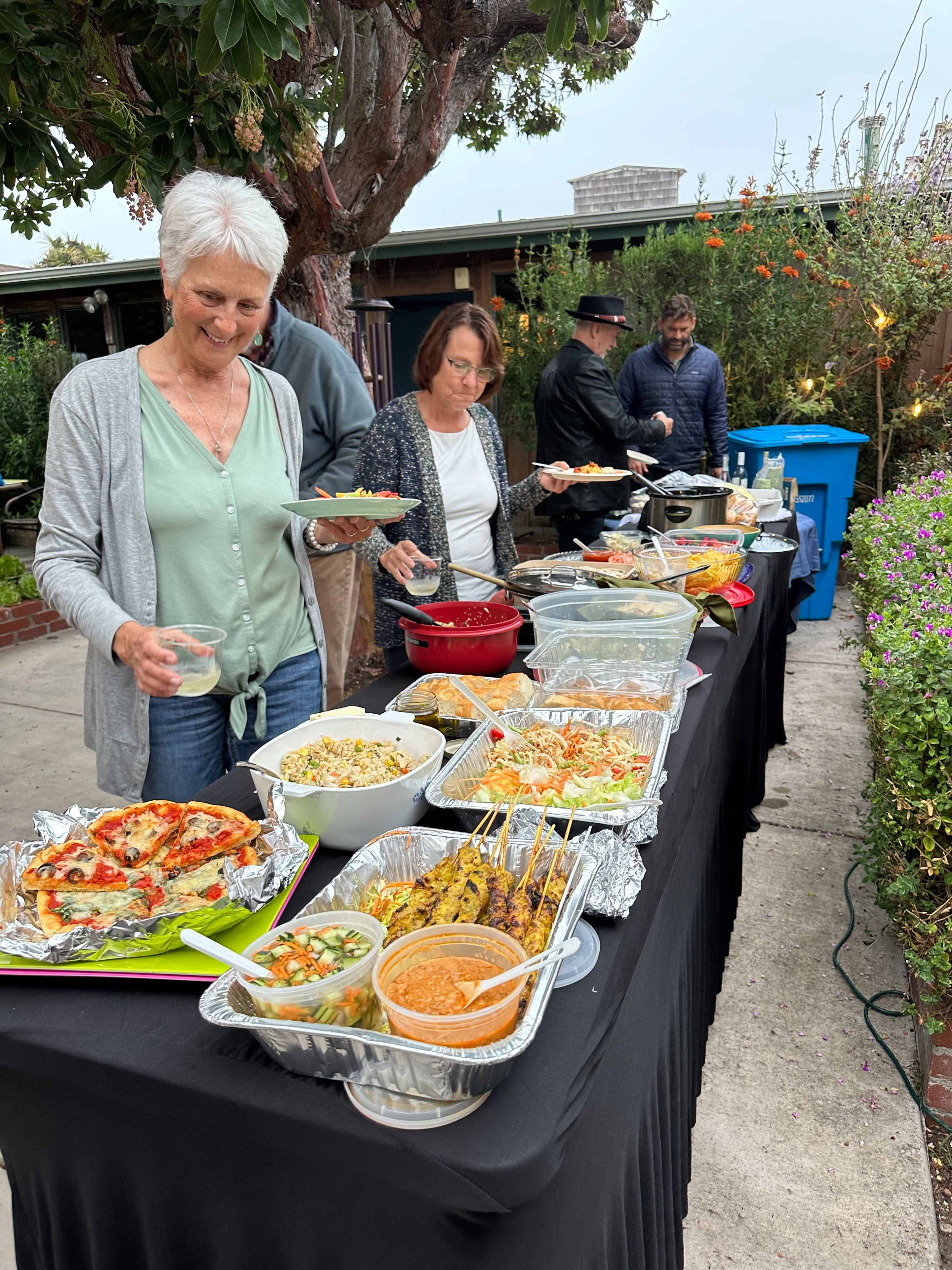 A group of people serves various dishes from a buffet-style table set outdoors, surrounded by greenery and blooming flowers.