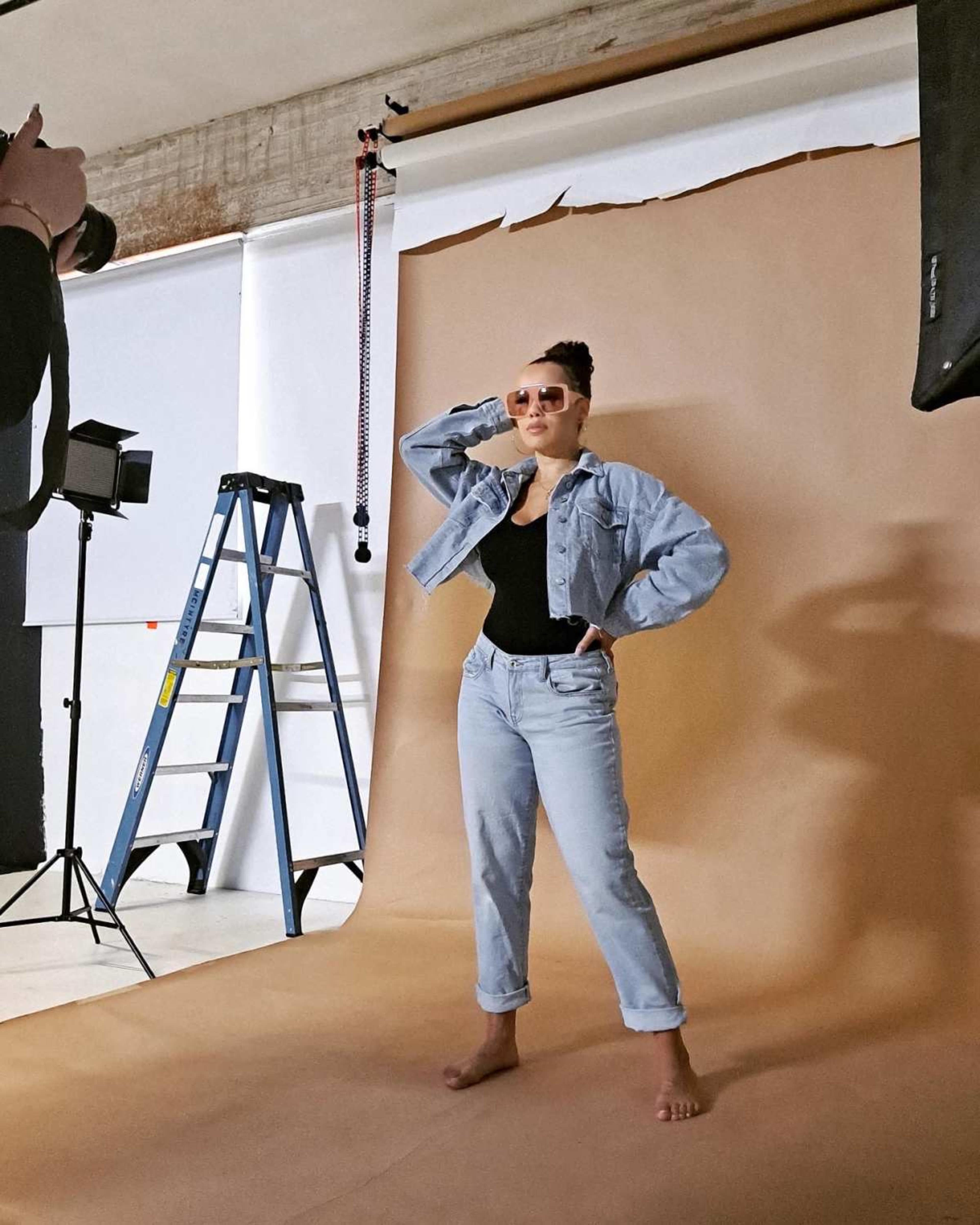 A model poses in a denim jacket and jeans against a beige backdrop in a photography studio.