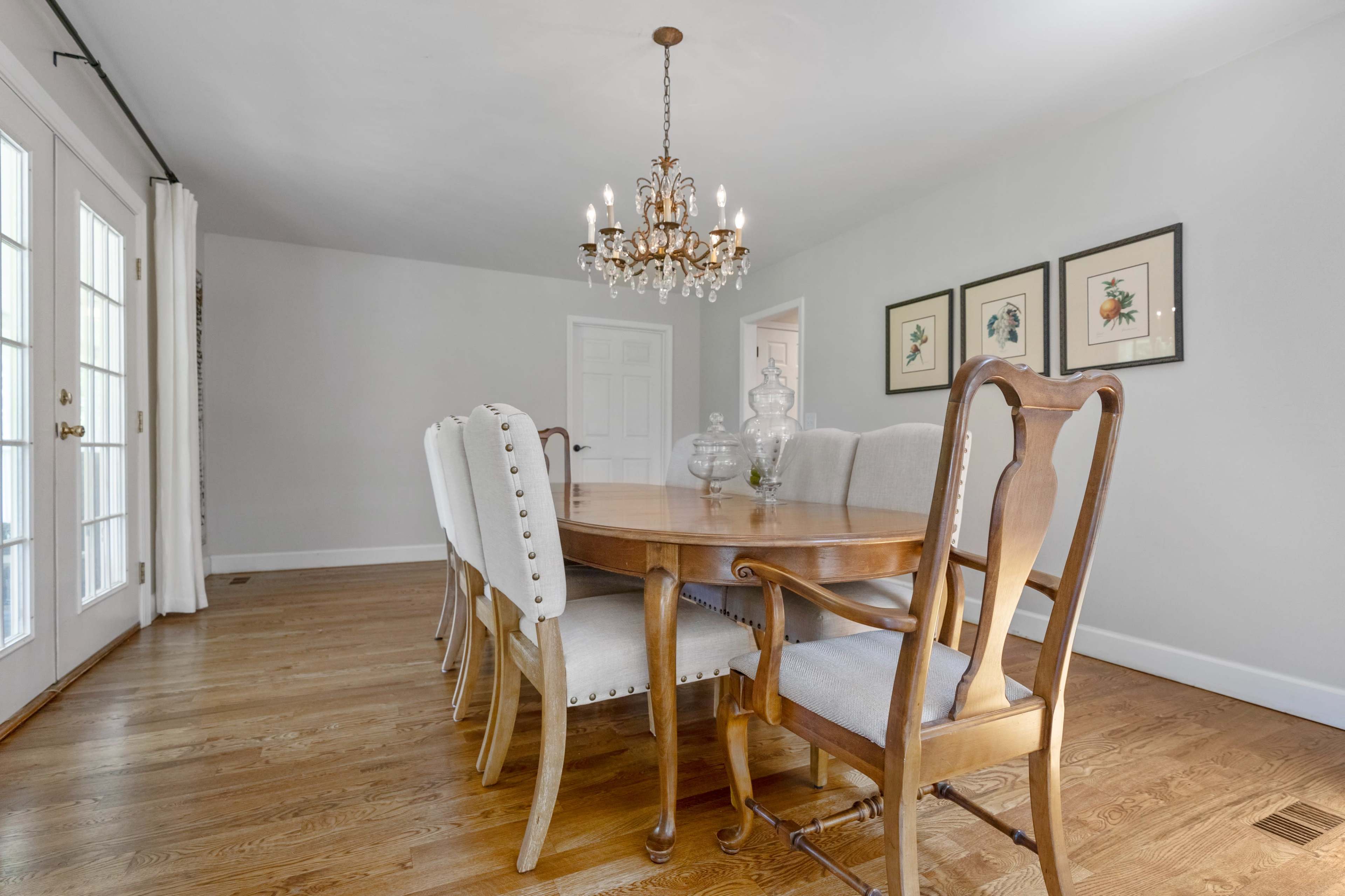 A wooden dining table with upholstered chairs is positioned under a chandelier in a well-lit dining room featuring light gray walls and hardwood flooring.
