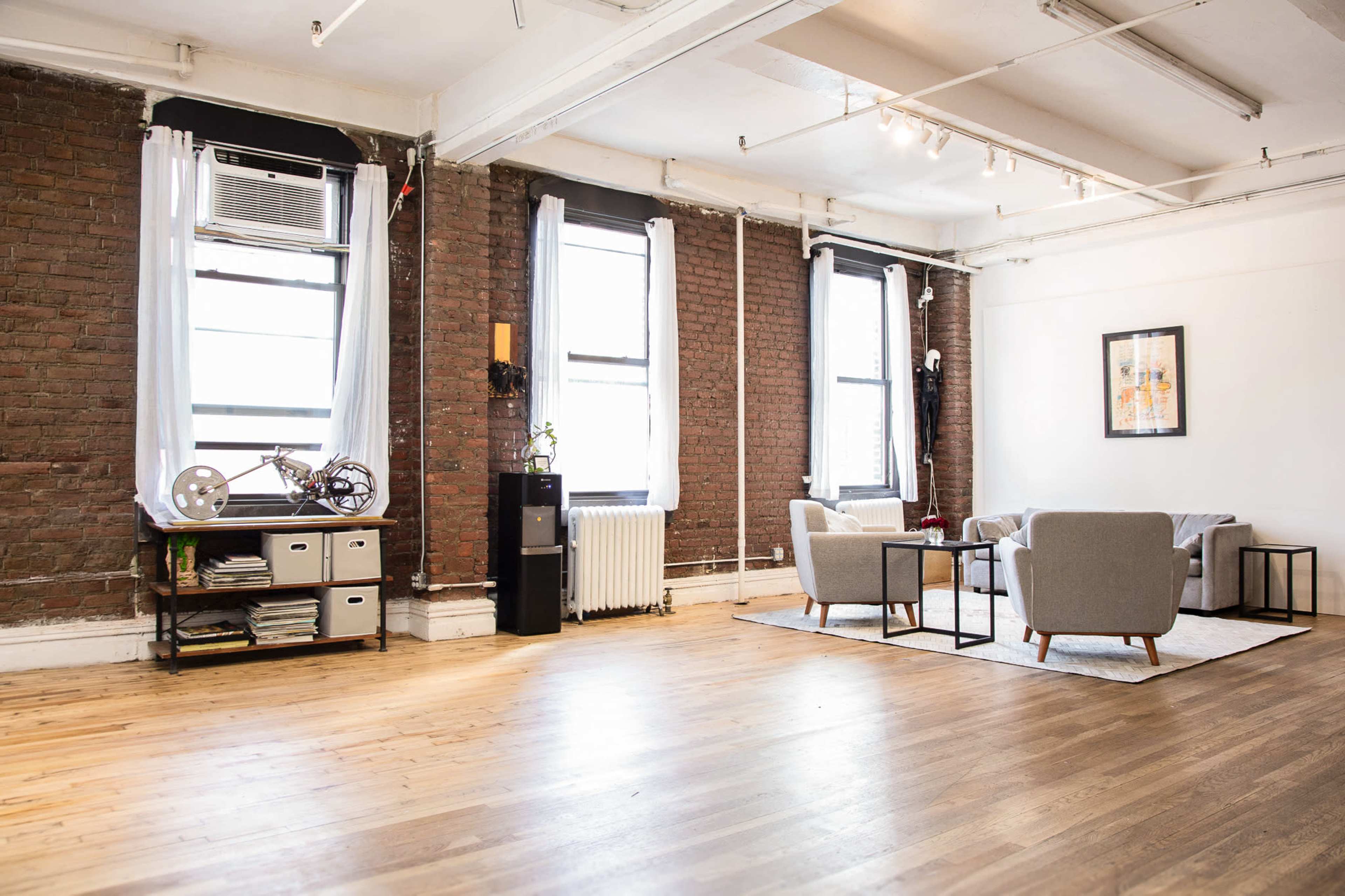 The image shows a spacious room with exposed brick walls, large windows, hardwood floors, and minimal furnishings including two gray armchairs, a coffee table, and a storage unit.