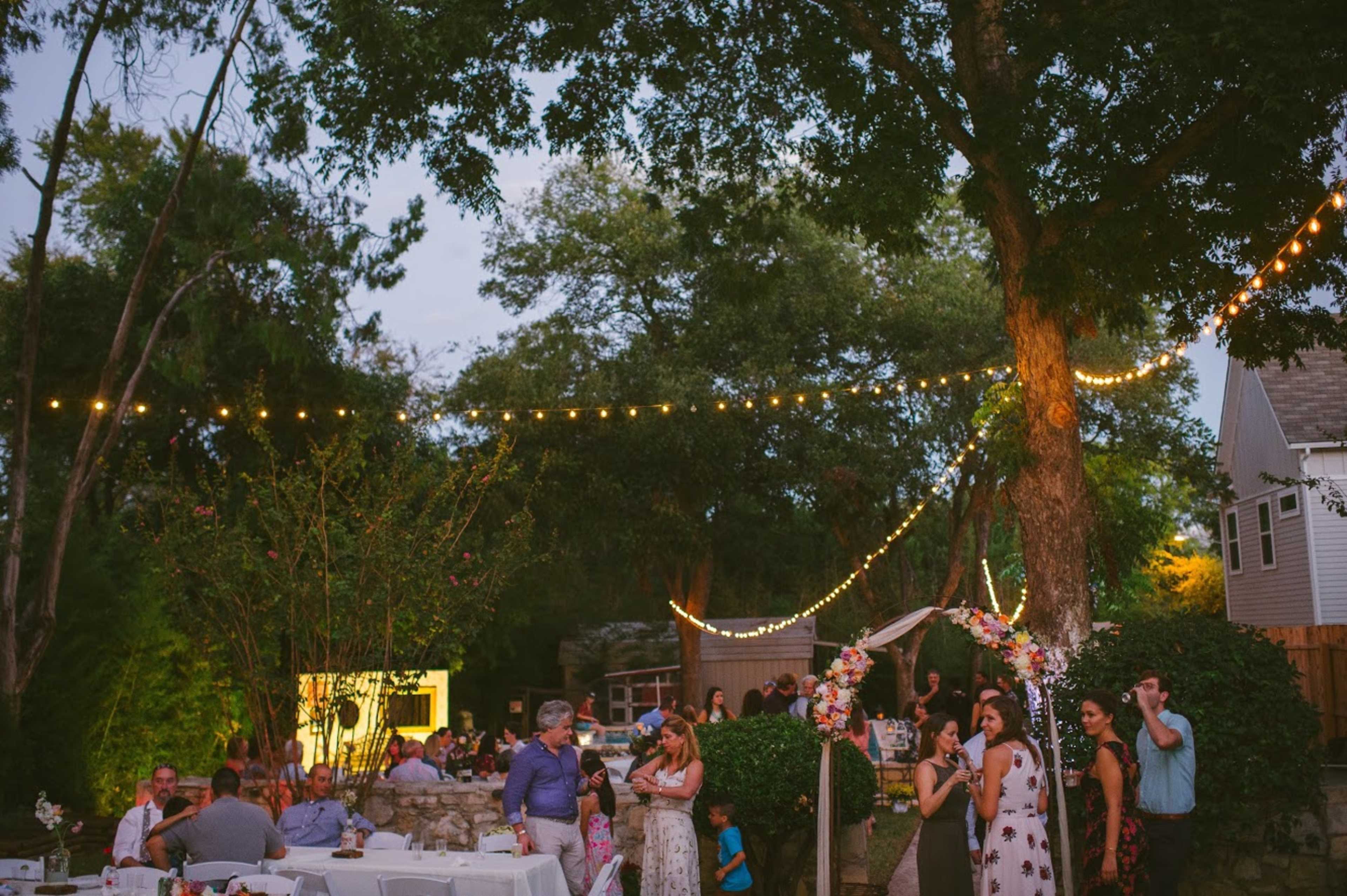 A gathering takes place in a backyard decorated with string lights, featuring guests socializing and enjoying the evening ambiance.